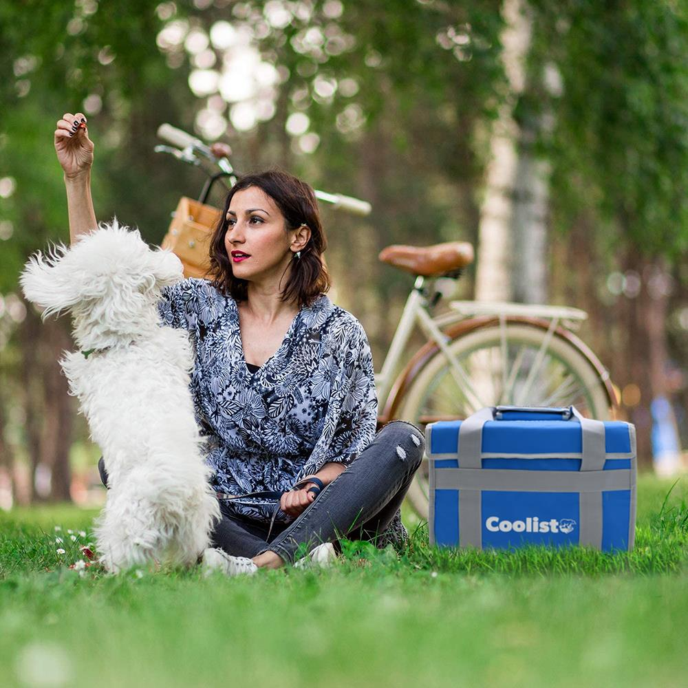 Bir kadın, beyaz bir köpekle oynarken çimlerde oturuyor, yanında kamp tipi buzdolabı ve bisiklet var.