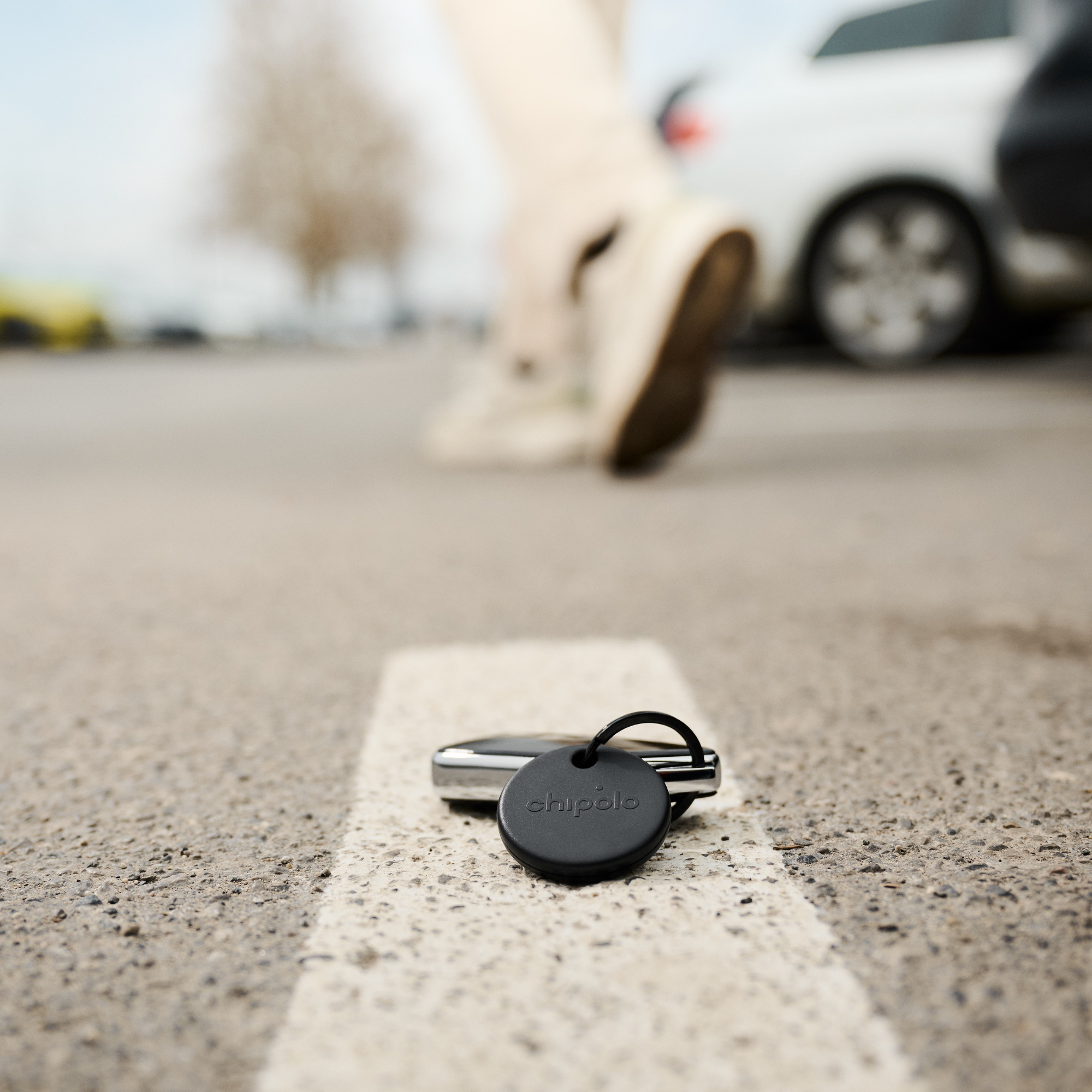 A black Chipolo key finder lies on a parking lot next to a car.