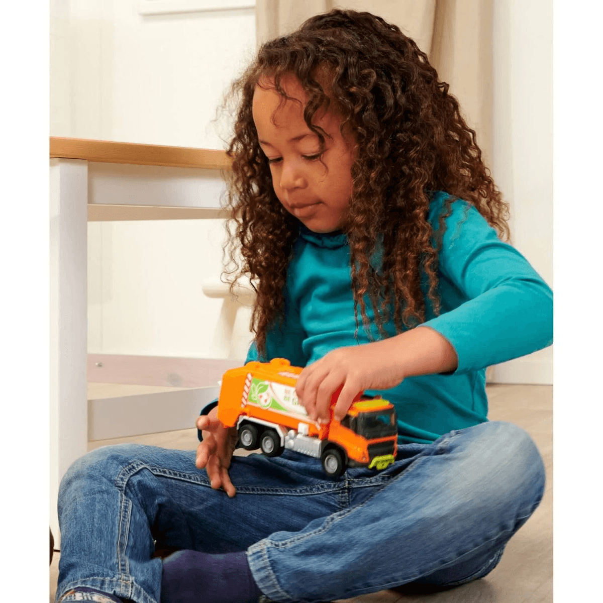 Ein kleines Mädchen mit lockigem Haar spielt mit einem orangefarbenen Müllwagen-Spielzeug.