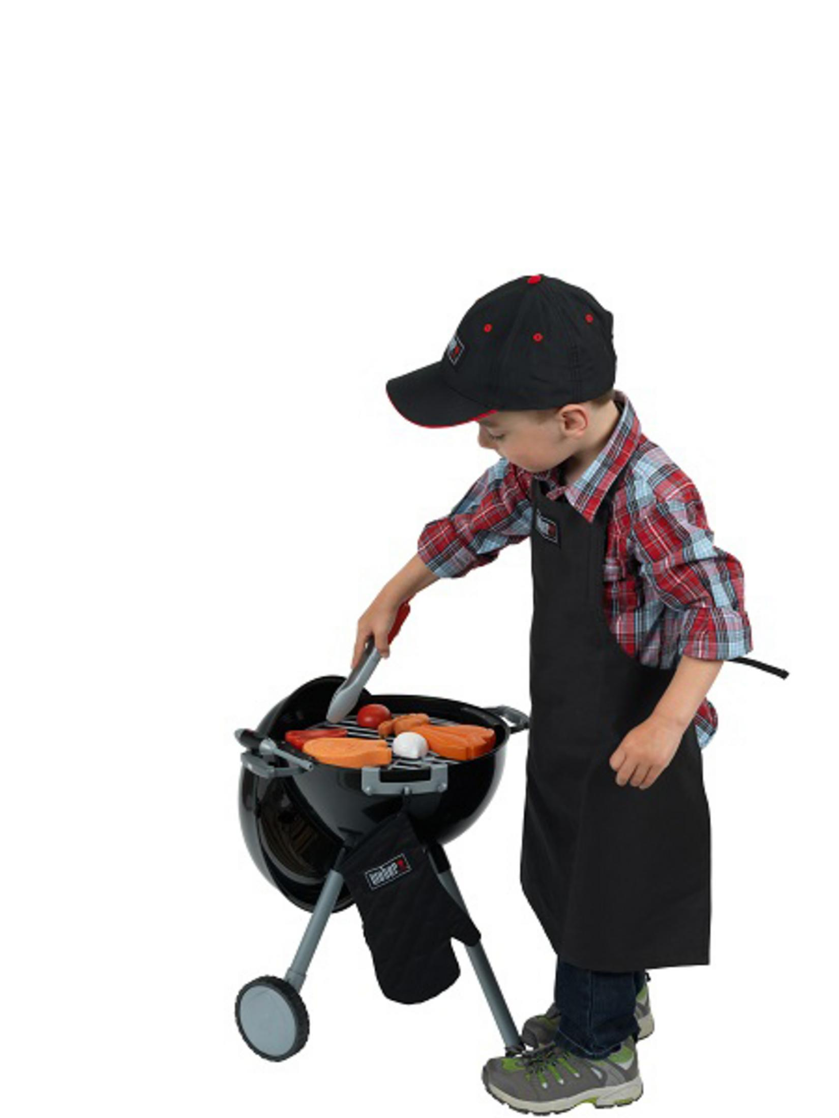 Un bambino con grembiule e cappellino griglia cibo giocattolo su un barbecue giocattolo.