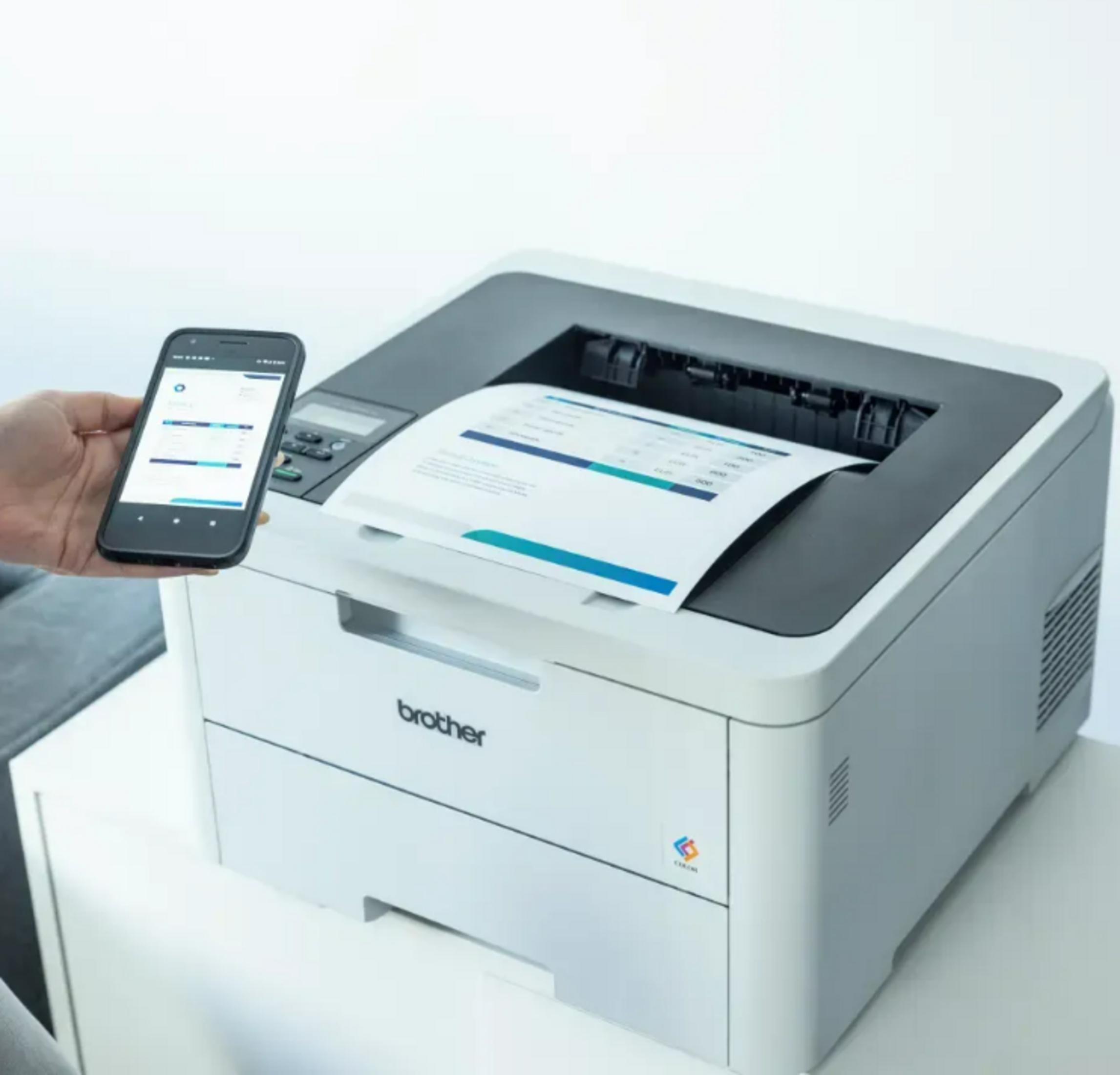 A smartphone is held over a Brother printer printing a sheet of paper.