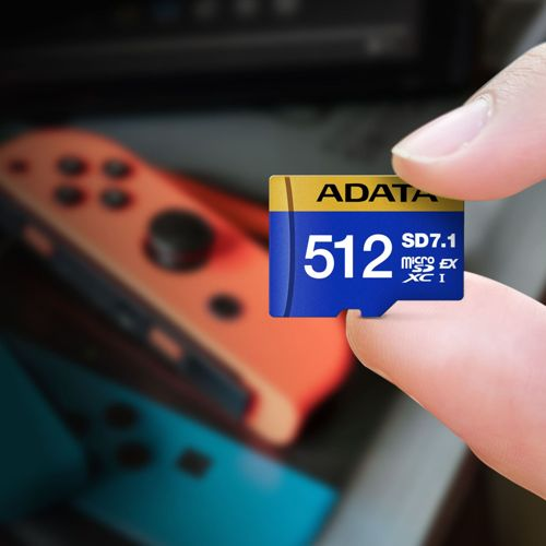 Un doigt tient une carte microSD ADATA de 512 Go. Une manette de jeu est floue en arrière-plan.