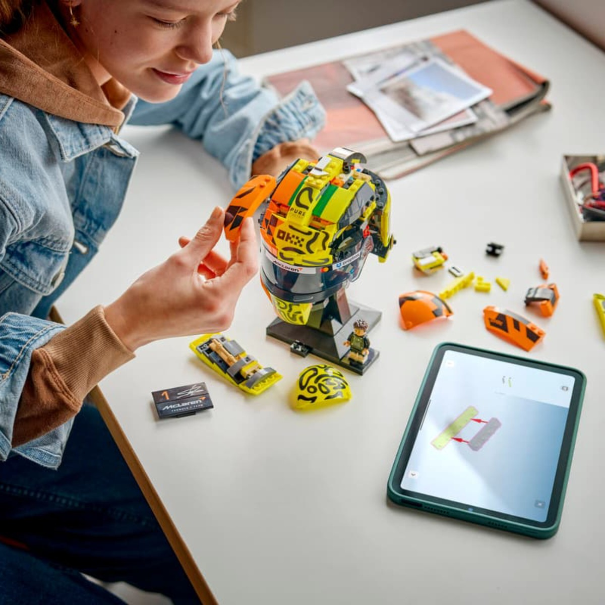 Kız, tablet talimatlarıyla sarı McLaren kaskı LEGO seti inşa ediyor. Parçalar etrafa saçılmış.