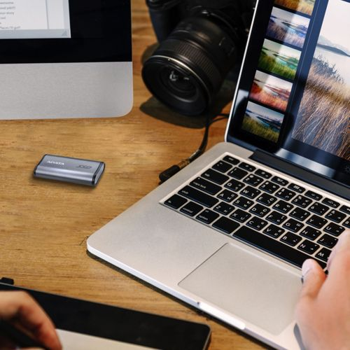 Chiavetta USB argento accanto a laptop, fotocamera e monitor su tavolo legno.