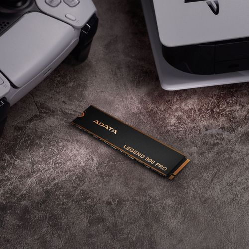Un SSD ADATA Legend 900 Pro nero è su una superficie scura e ruvida, vicino a un controller e console di gioco bianchi.
