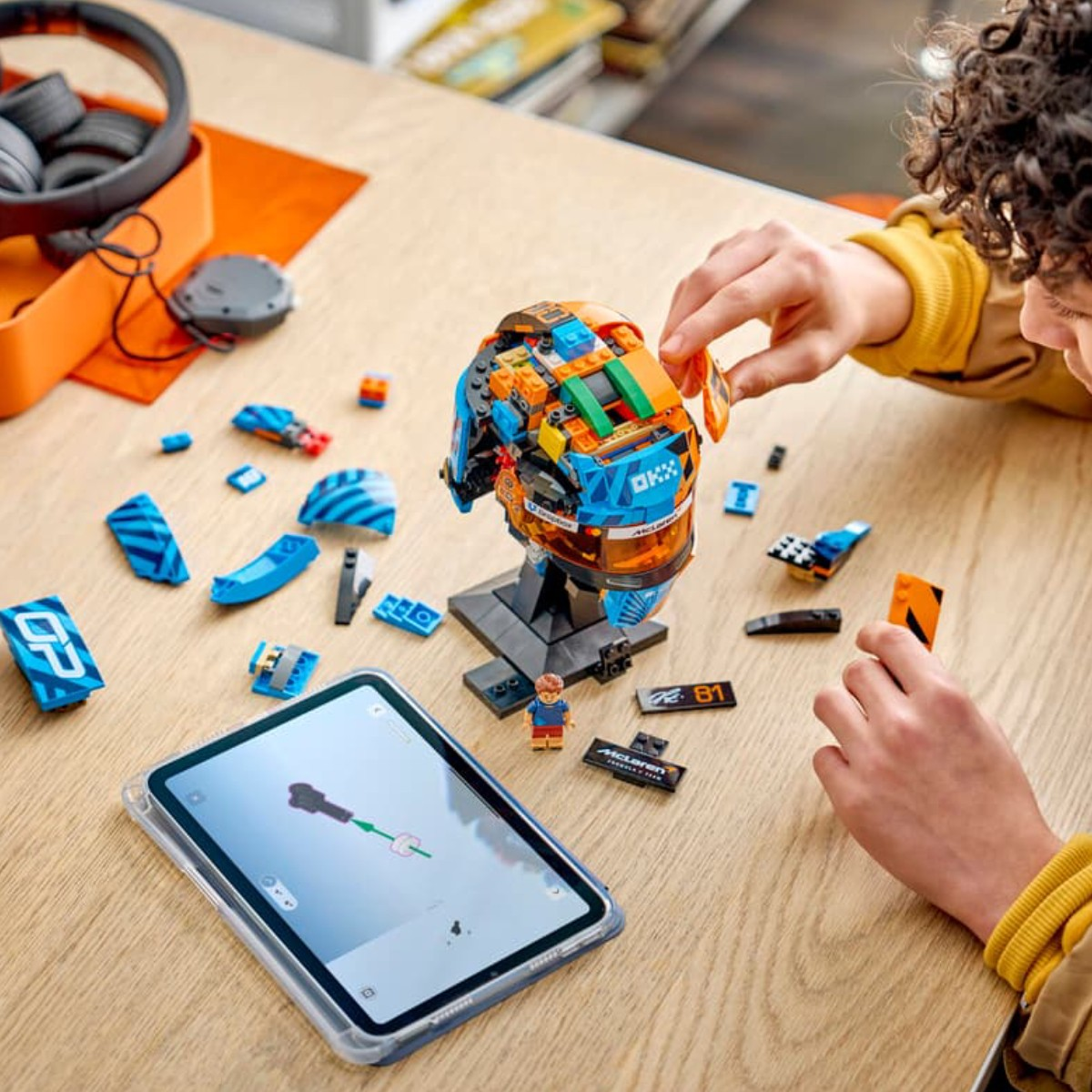 Çocuk, talimatları gösteren bir tabletle, bir stand üzerinde mavi ve turuncu Lego kaskı inşa ediyor.