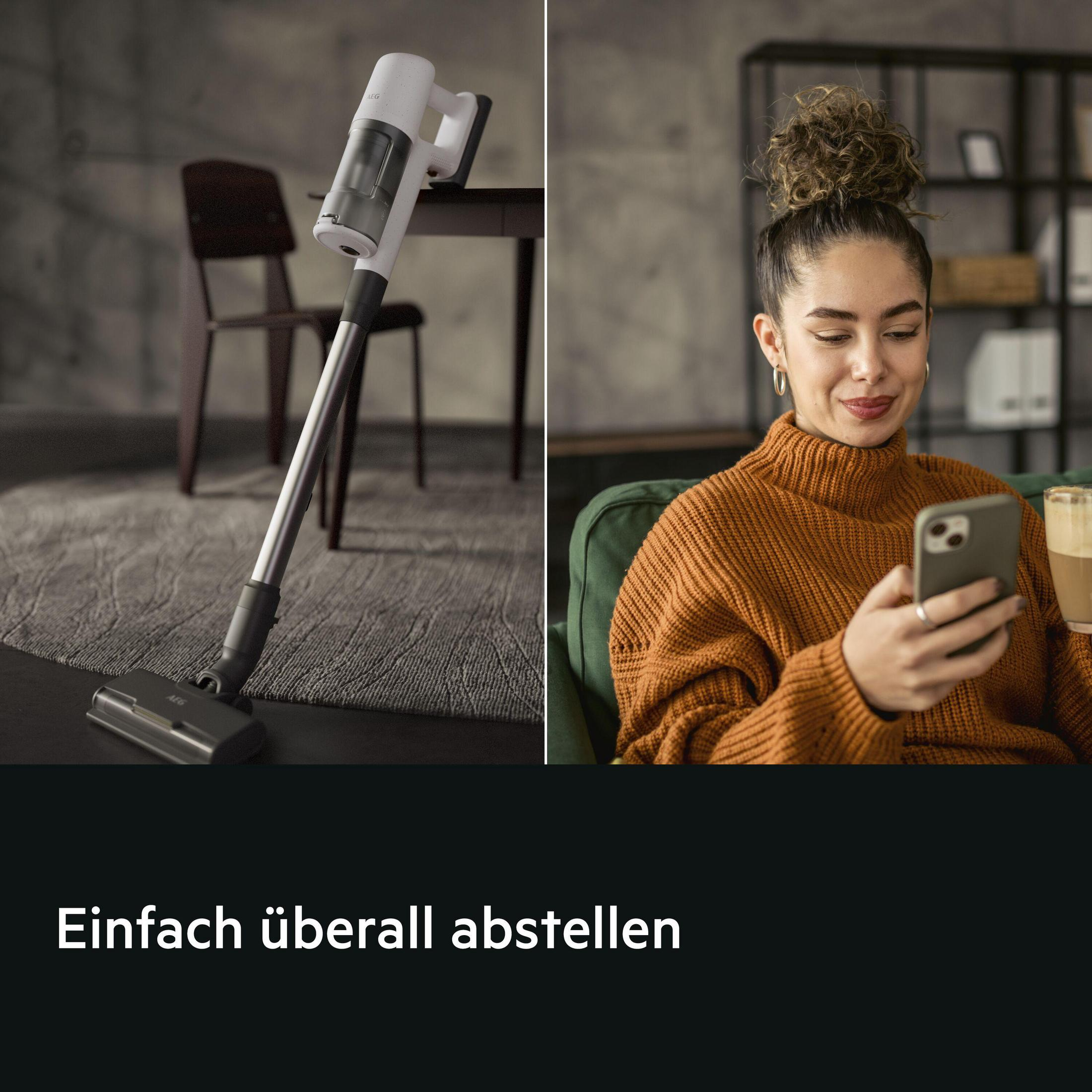 Ein weißer Staubsauger steht neben einem Tisch; eine Frau hält ein Handy.