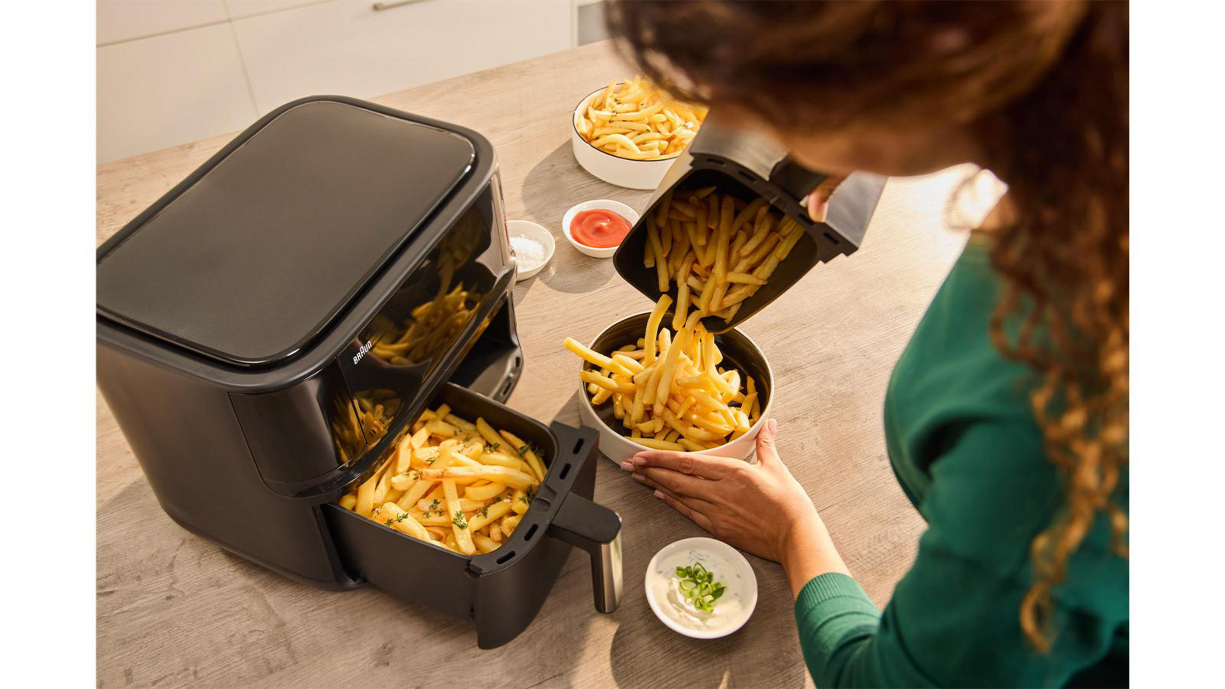 Eine Frau schüttet Pommes aus einem schwarzen Heißluftfritteusenkorb in eine Schüssel.