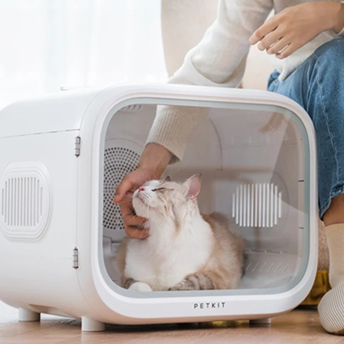 Una persona accarezza un gatto all'interno di un'asciugatrice bianca per animali con finestra trasparente.