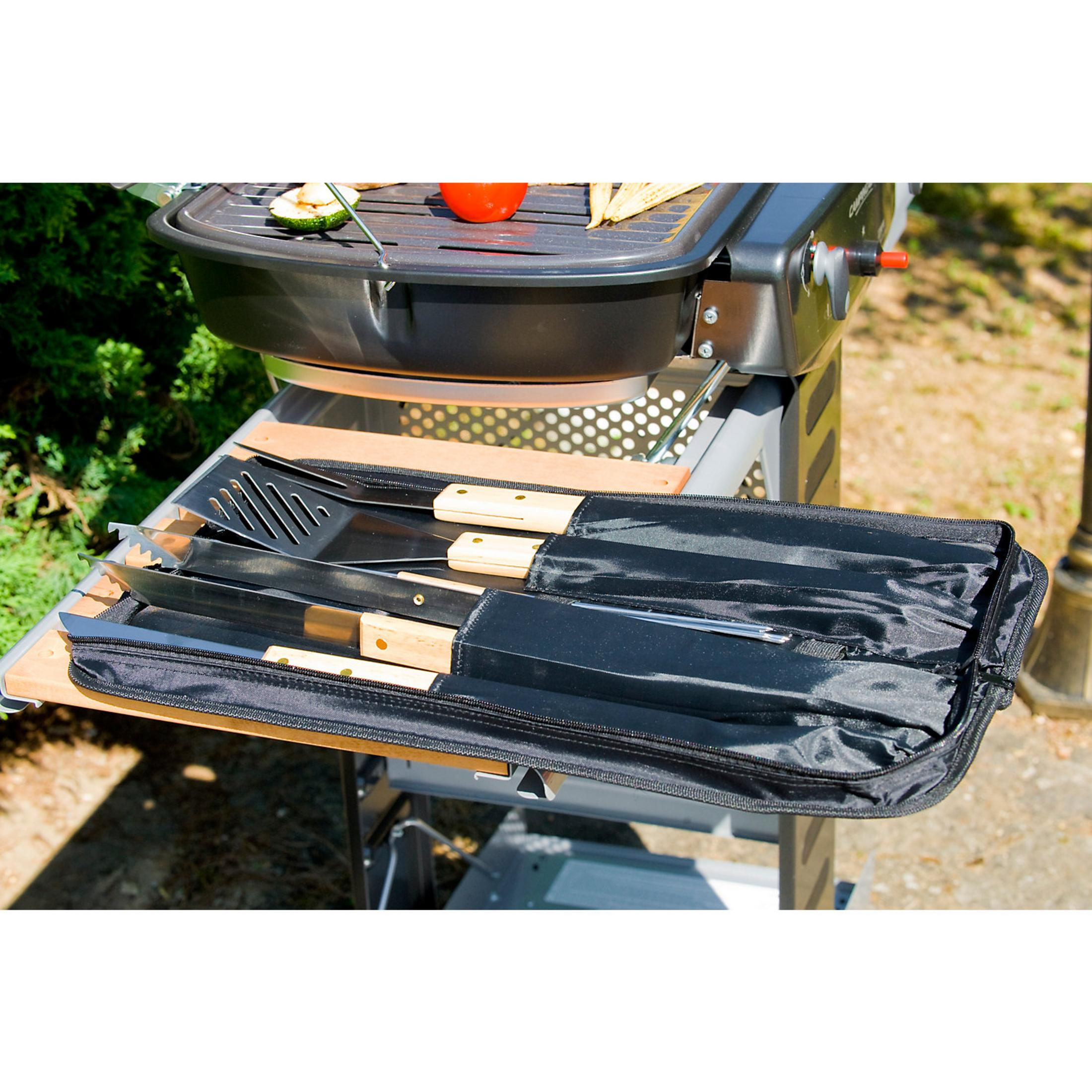Pinzas para parrilla y utensilios de cocina con mangos de madera en un estuche negro junto a la parrilla.