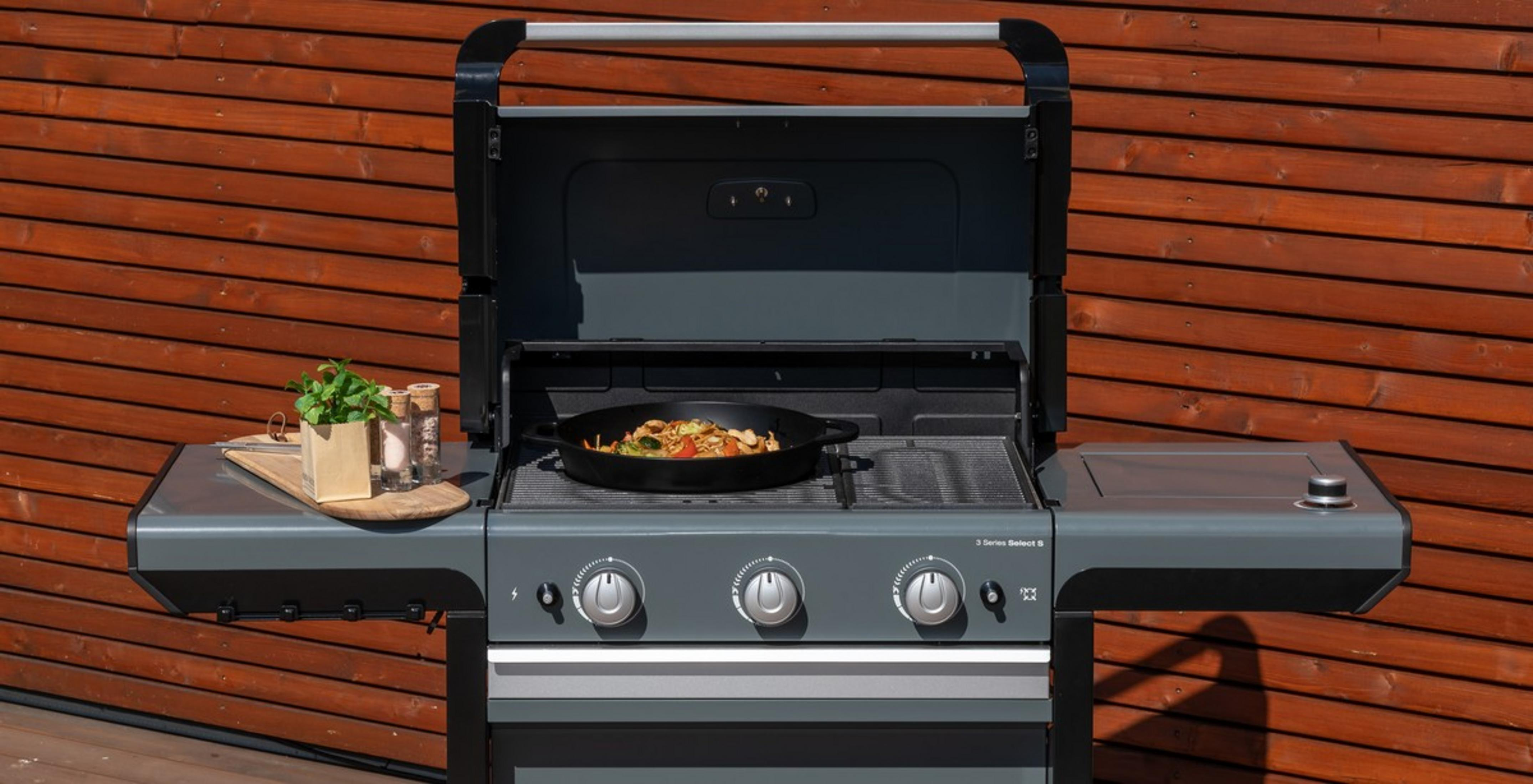 Una parrilla de gas con tapa abierta está frente a una pared de madera. Un wok con comida está en la rejilla.