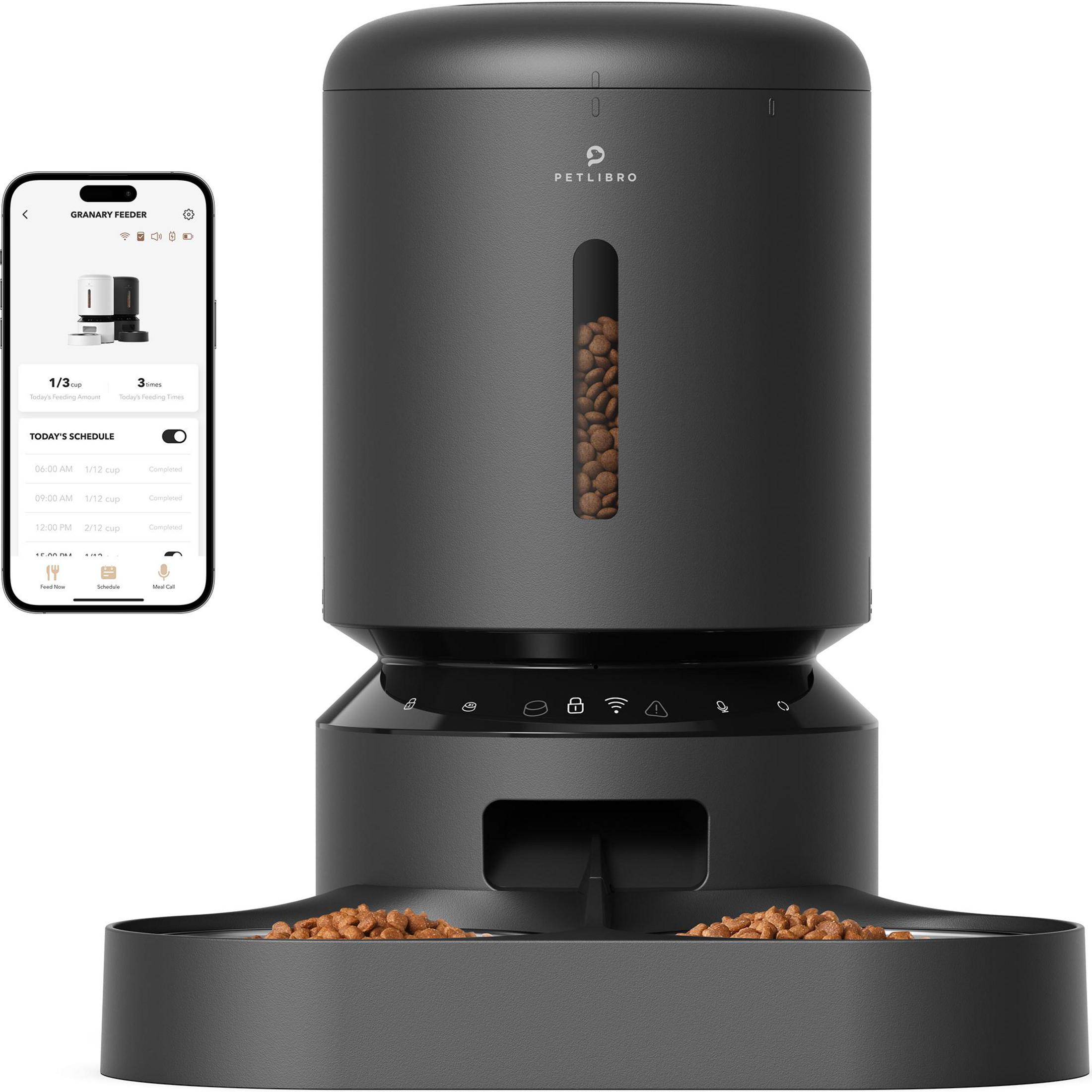 A black automatic pet feeder with a smartphone next to it showing the feeding schedule.
