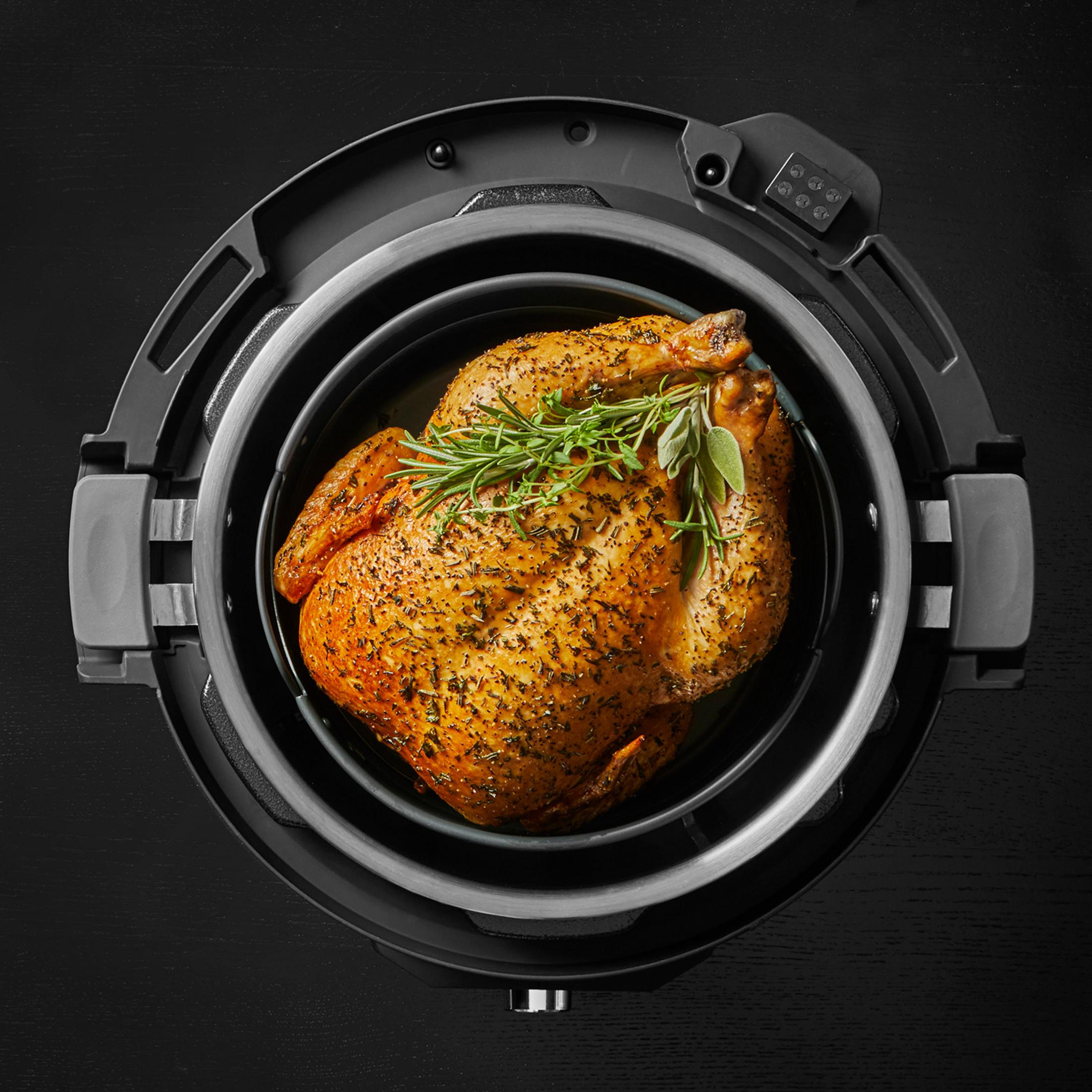 Een goudbruine gebraden kip met kruiden in een zwarte airfryer op een donkere achtergrond.