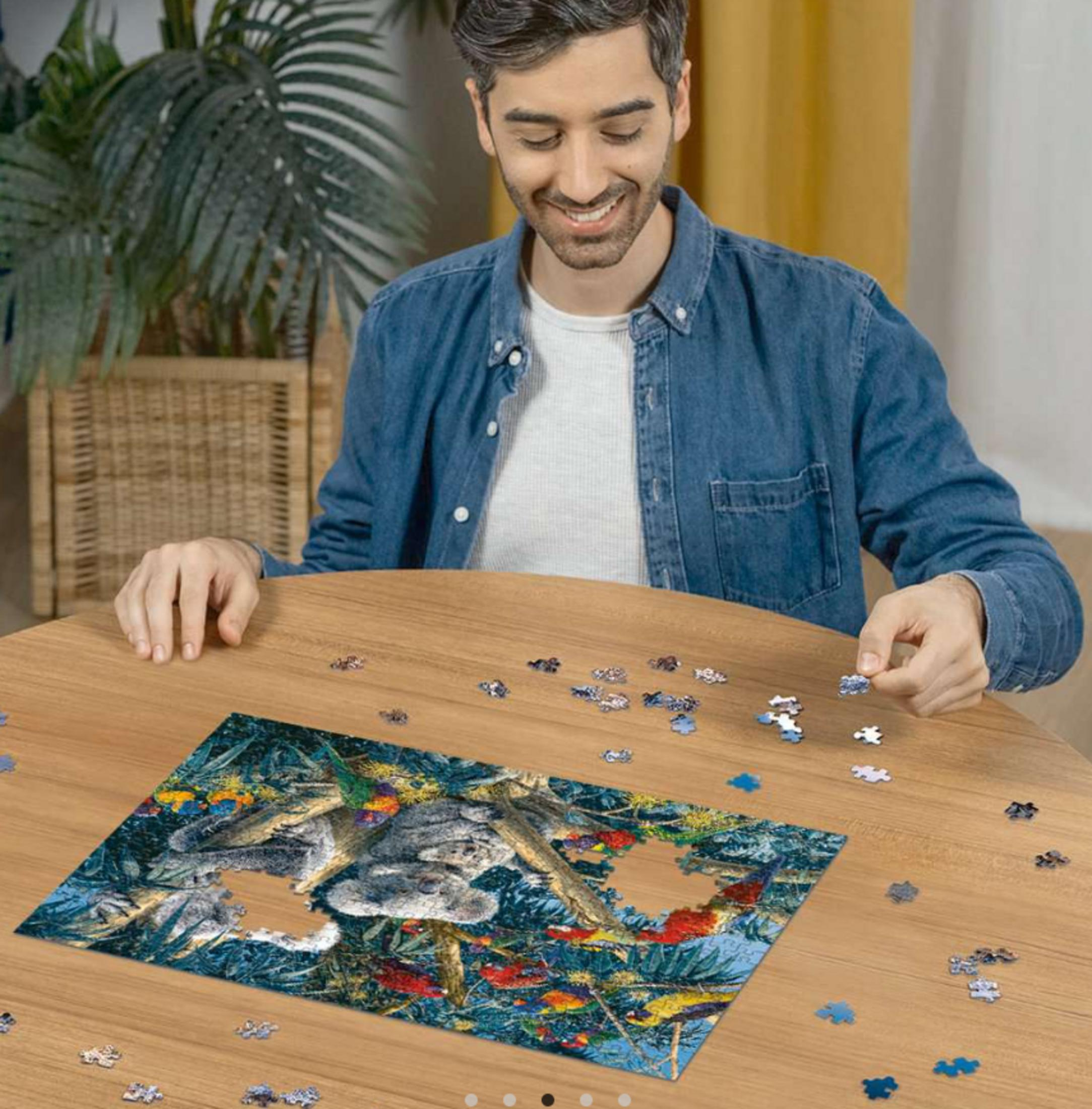 Mann im blauen Hemd puzzelt Koala und Papageienbild auf Holztisch.
