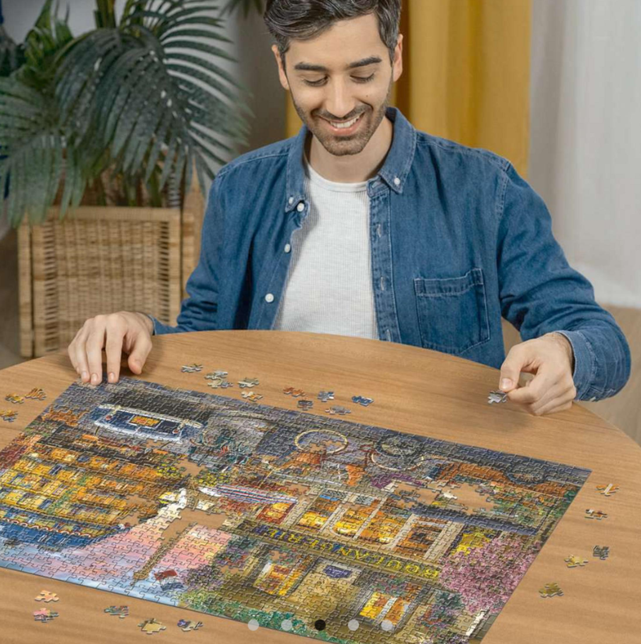 Un homme assis à une table en bois assemble un puzzle. Des plantes en arrière-plan.