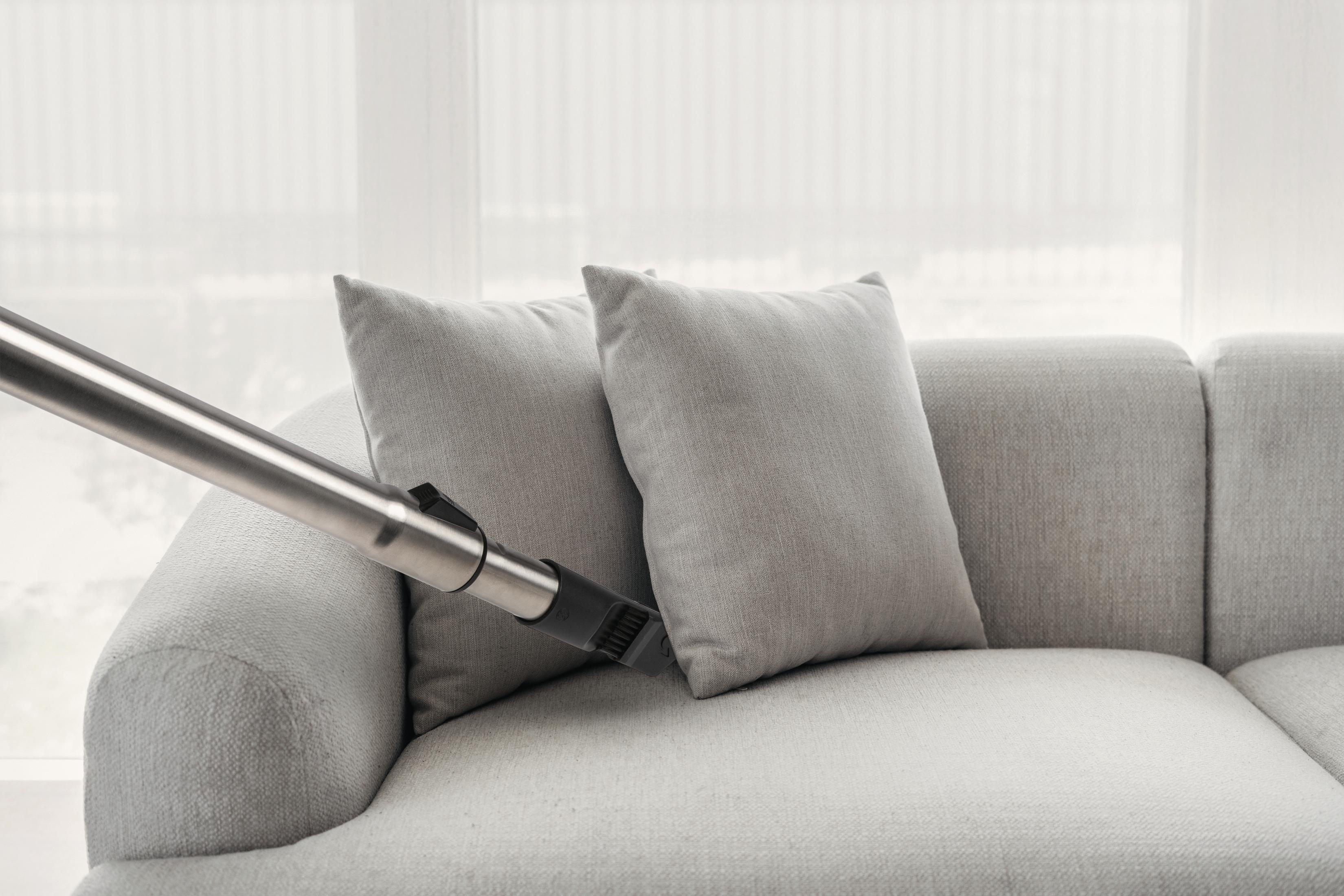 Ein Staubsaugerrohr reinigt ein graues Sofa mit zwei Kissen.