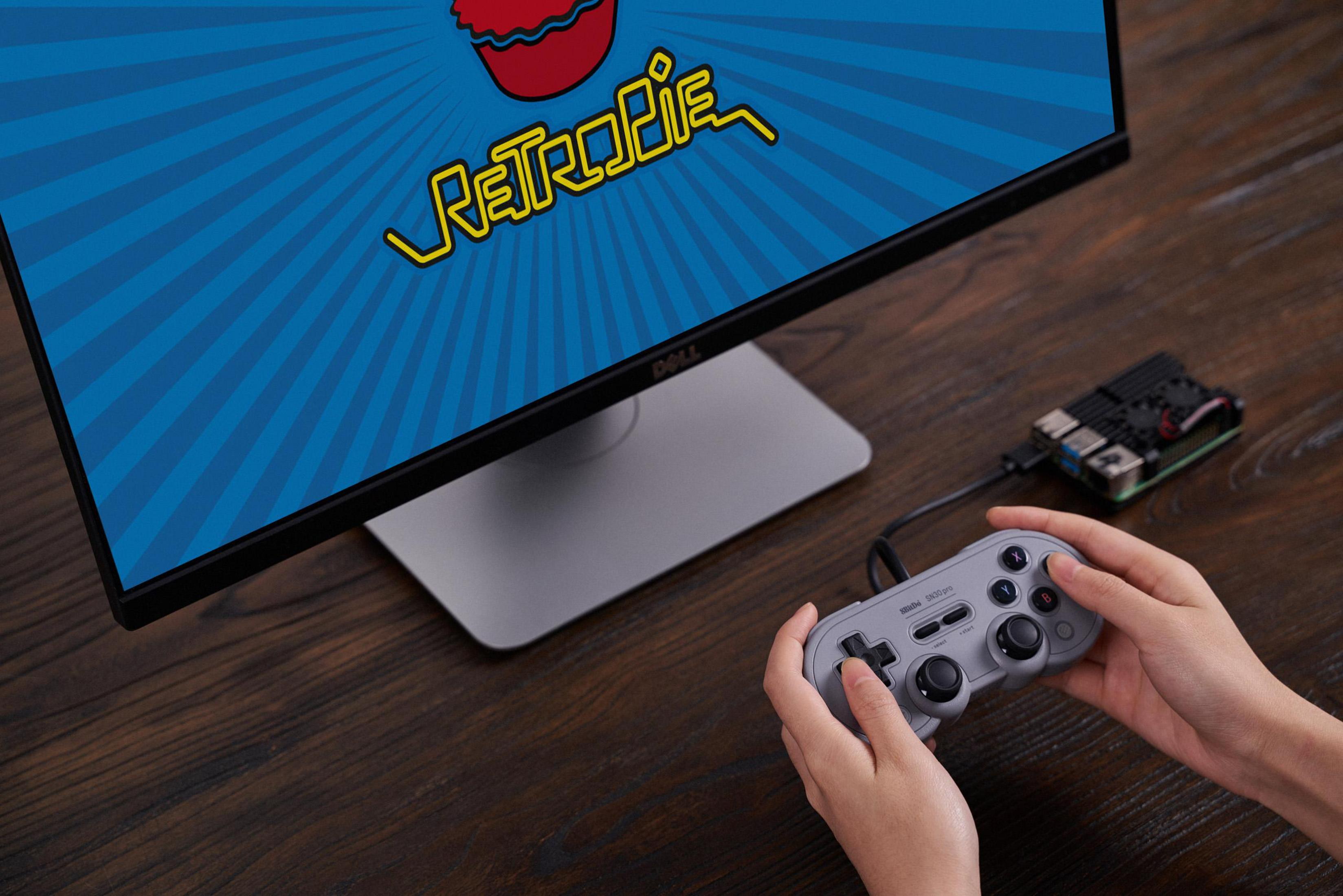 Mani tengono un gamepad grigio collegato a un Raspberry Pi, davanti a un monitor Dell che mostra Retropie.
