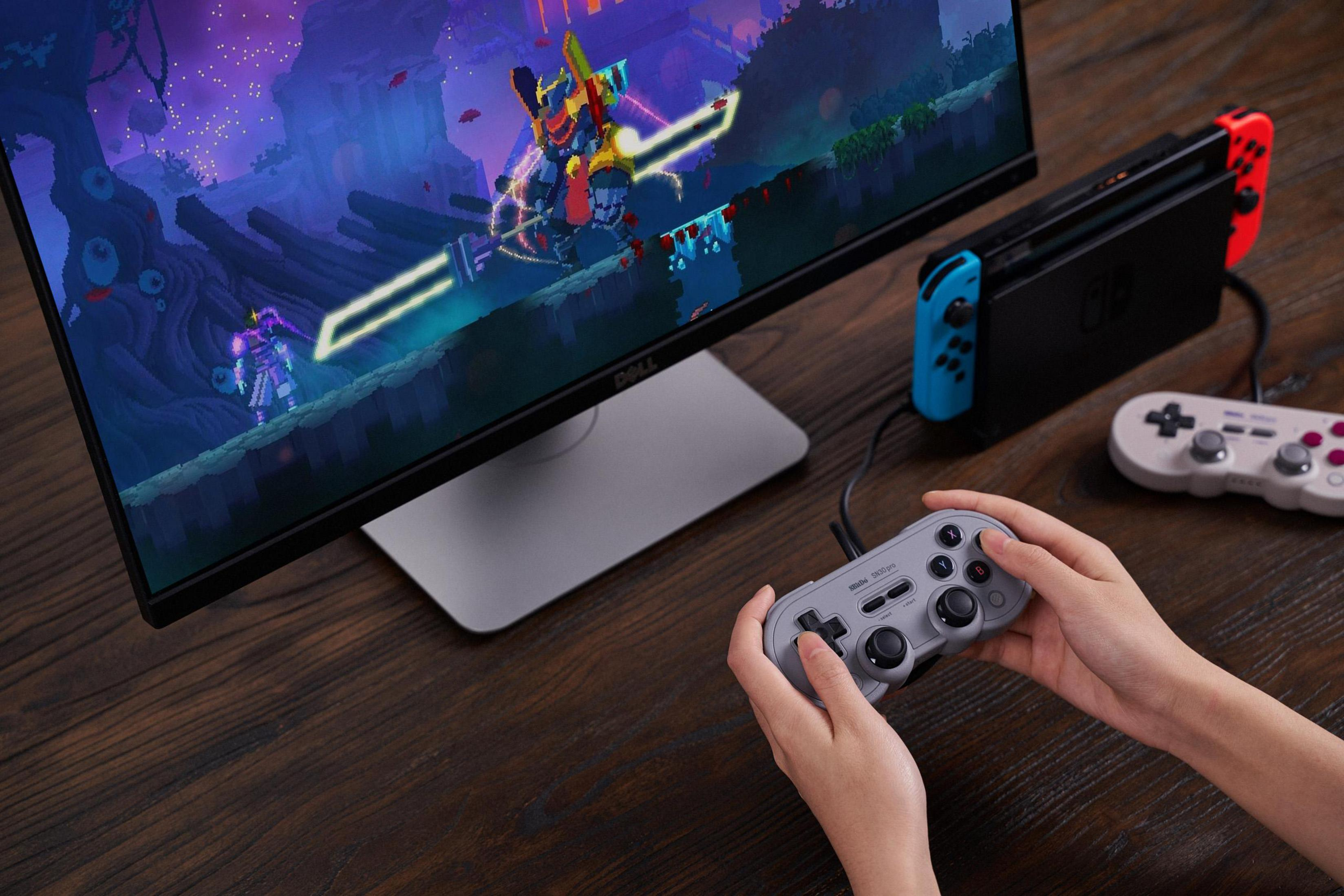 Una Nintendo Switch con controller e un giocatore tiene un controller grigio davanti a un monitor.
