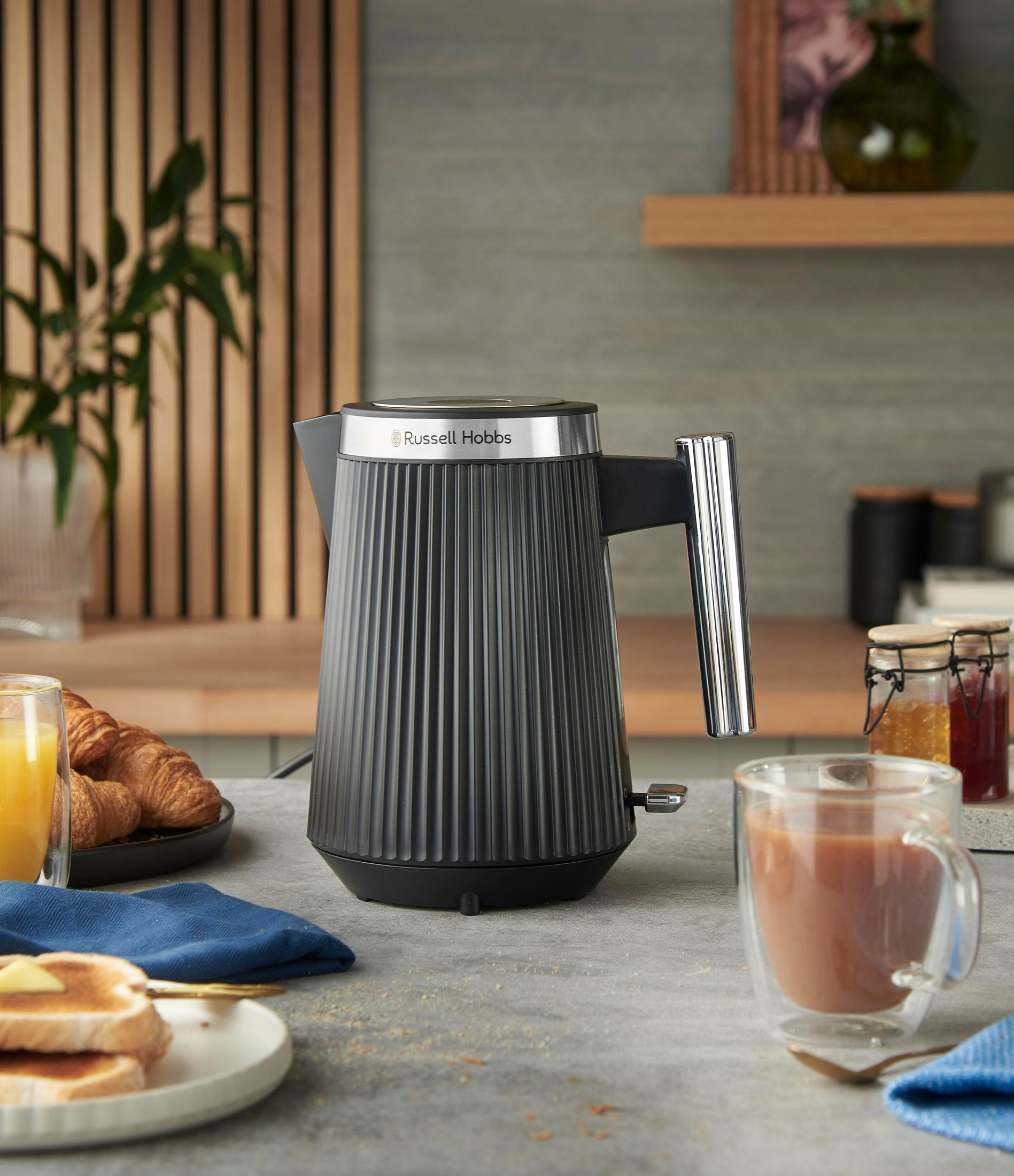 A black kettle sits on a table with toast, croissants, and juice.