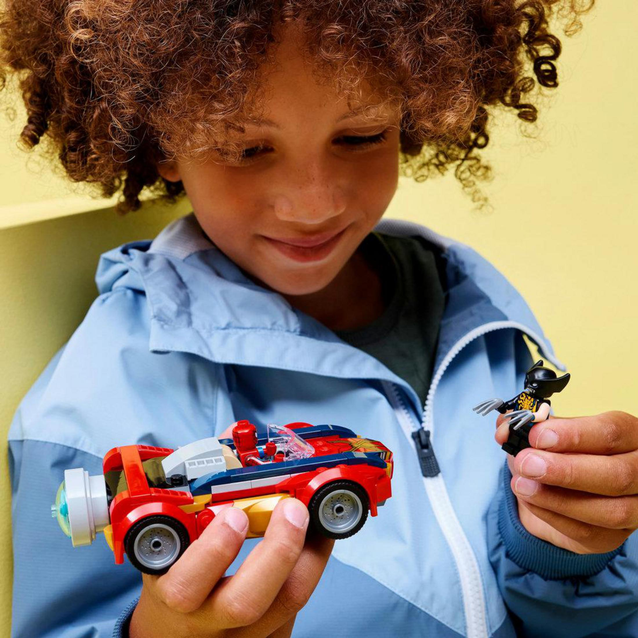 Un bambino con i capelli ricci tiene una macchinina Lego rossa e blu e una minifigure di Wolverine.