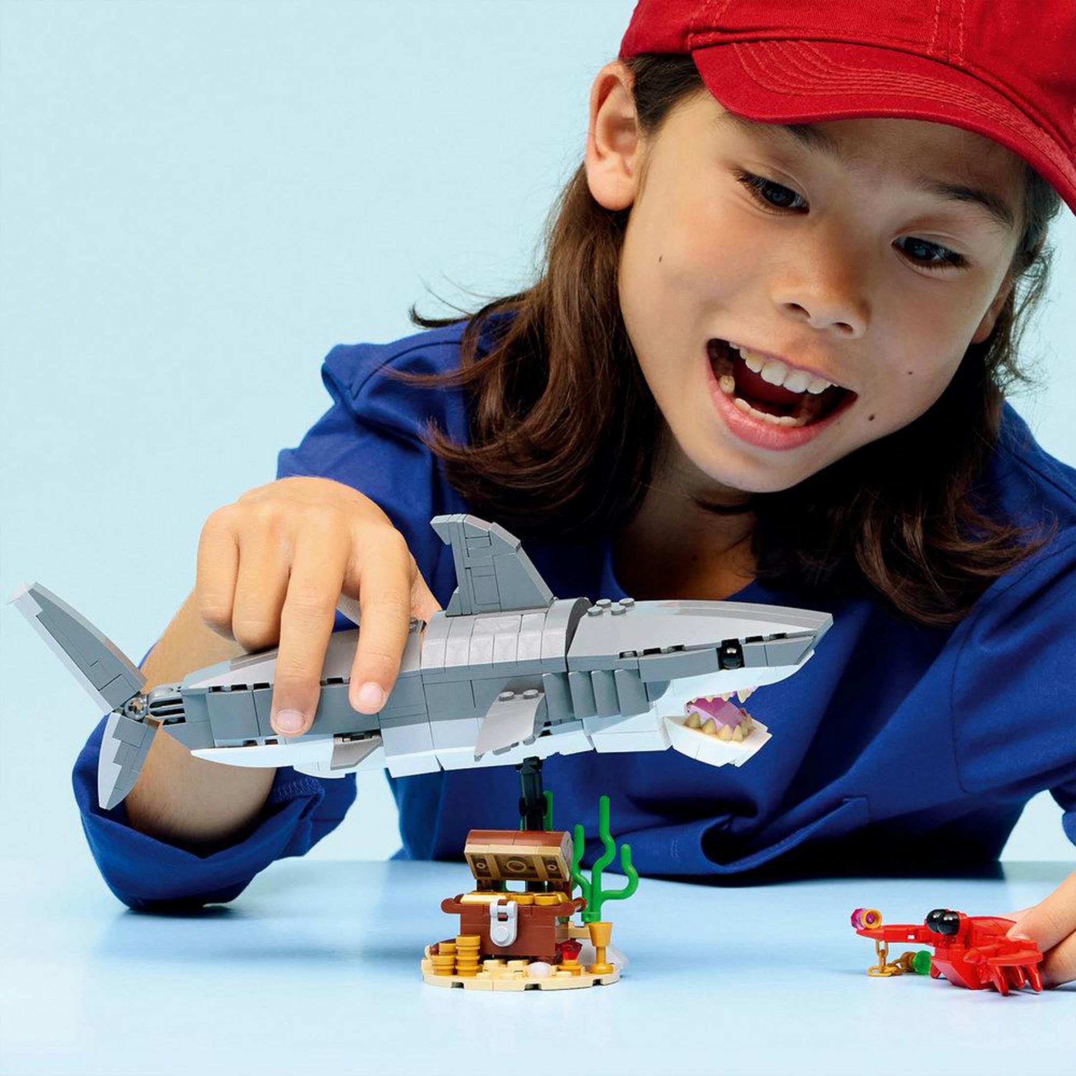Kırmızı şapkalı bir çocuk, hazine sandığının üzerinde bir Lego köpekbalığı tutuyor.