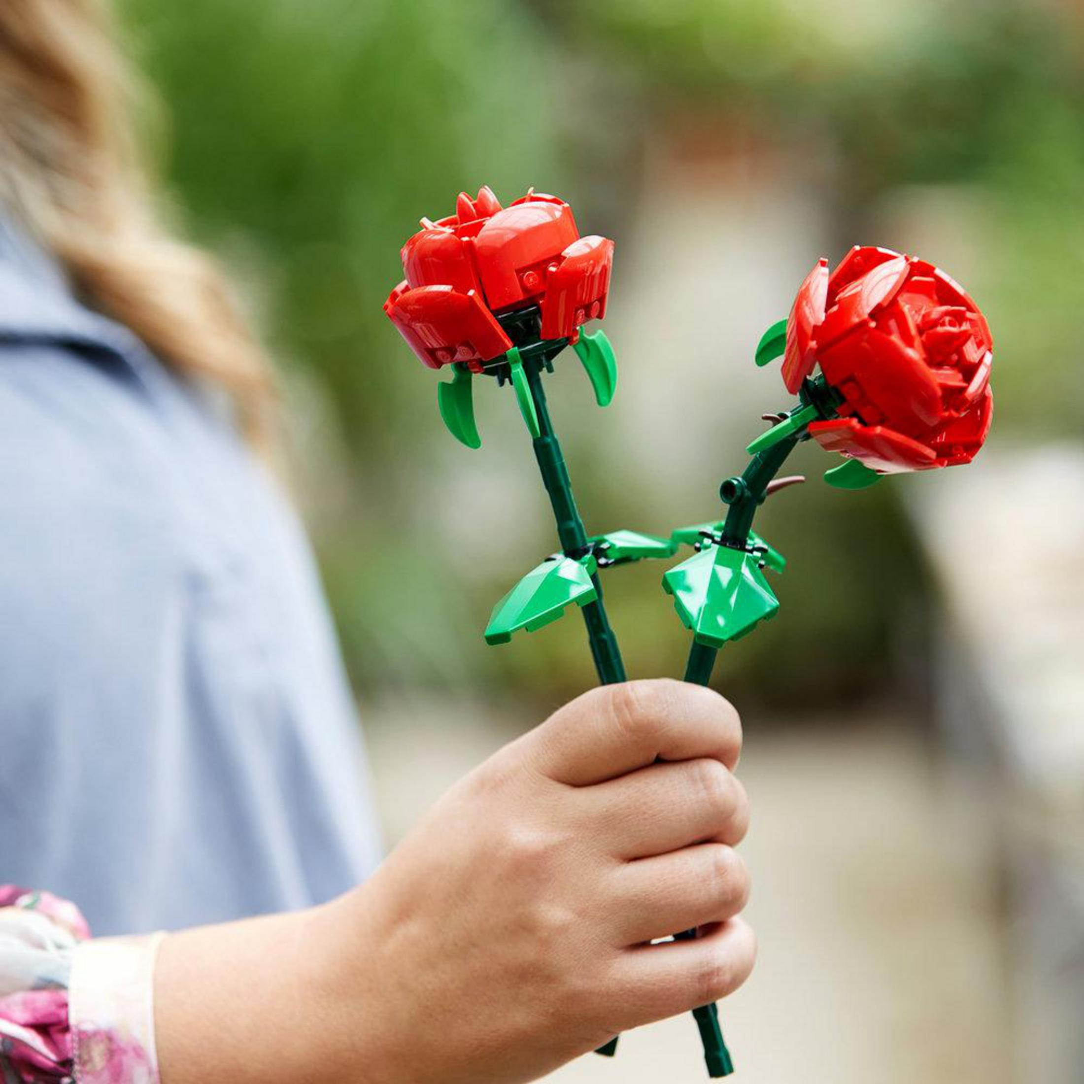 Persona sosteniendo dos rosas rojas de Lego con tallos y hojas verdes.