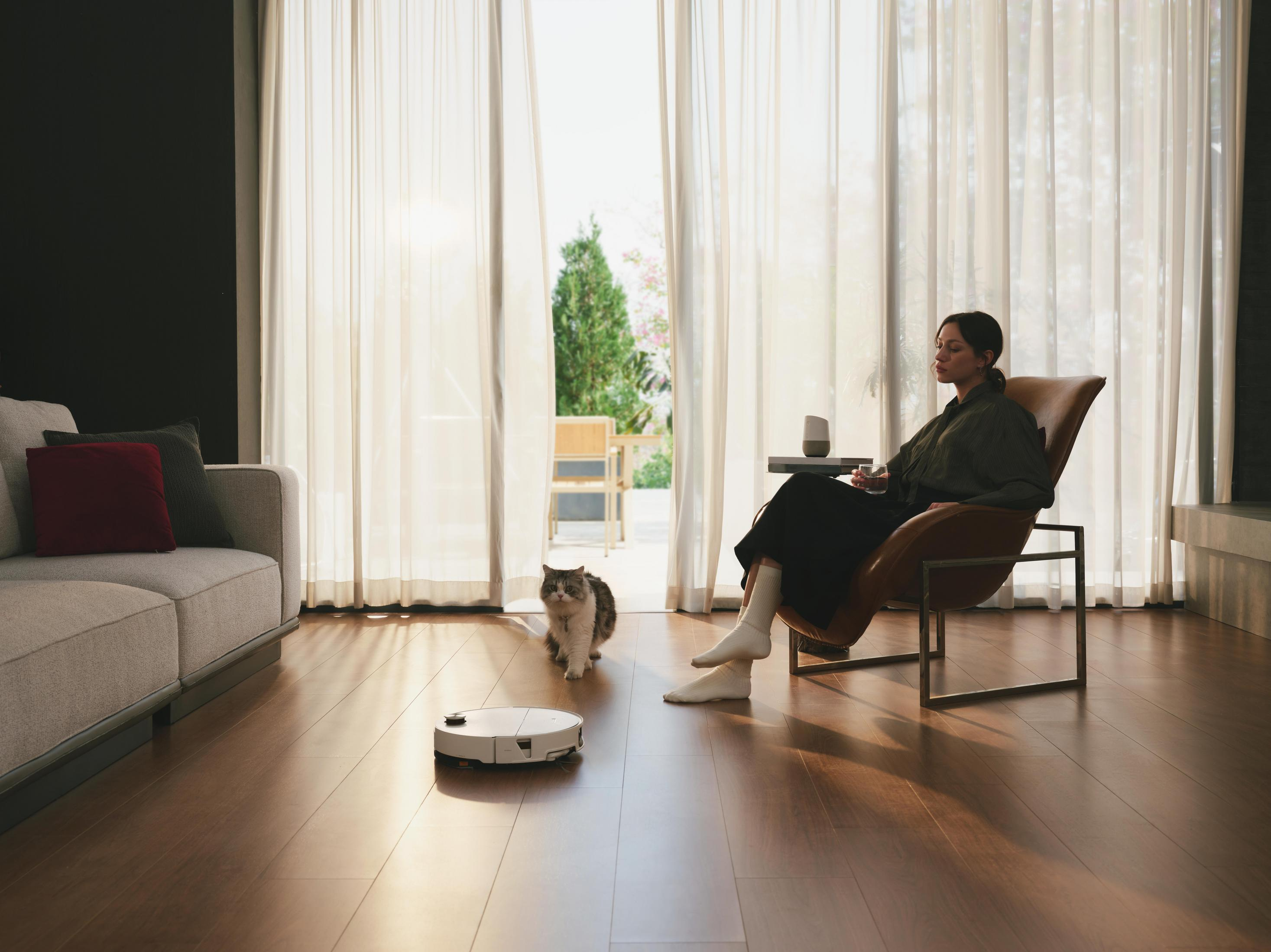 Eine Frau sitzt im Sessel, ein Staubsaugerroboter und eine Katze sind auf dem Holzboden.