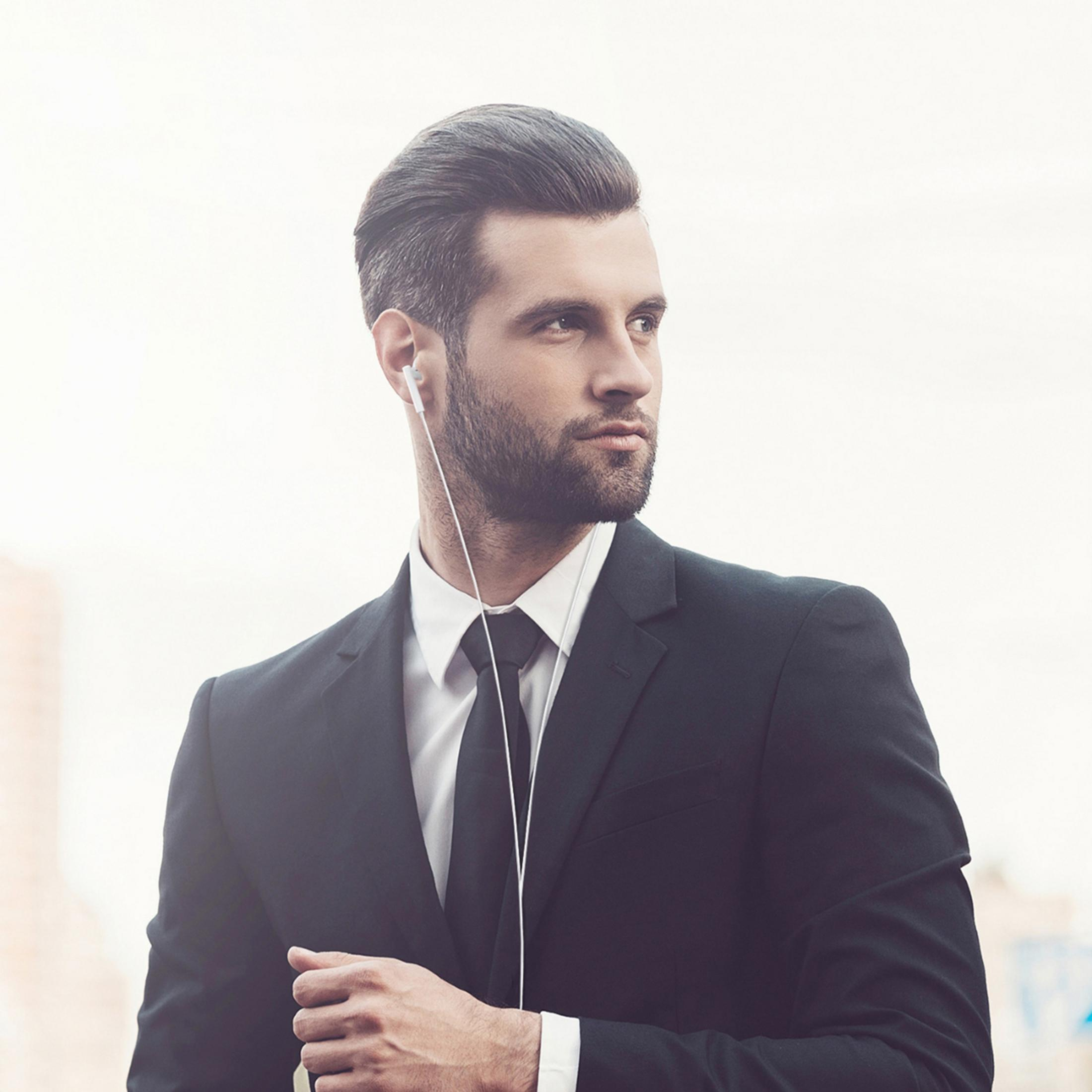 A man in a black suit wearing white earbuds looks to the right.