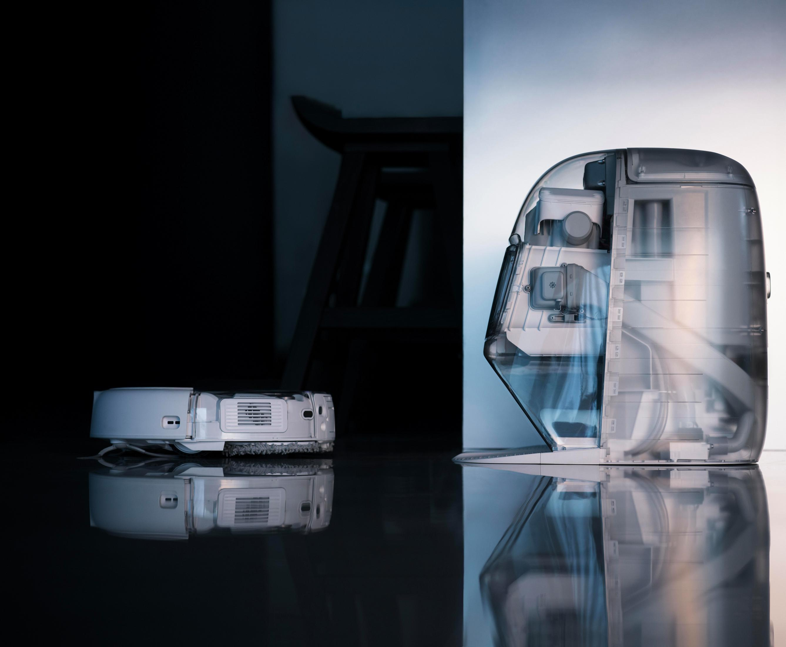 A white robot vacuum on a dark, reflective floor next to a transparent machine.