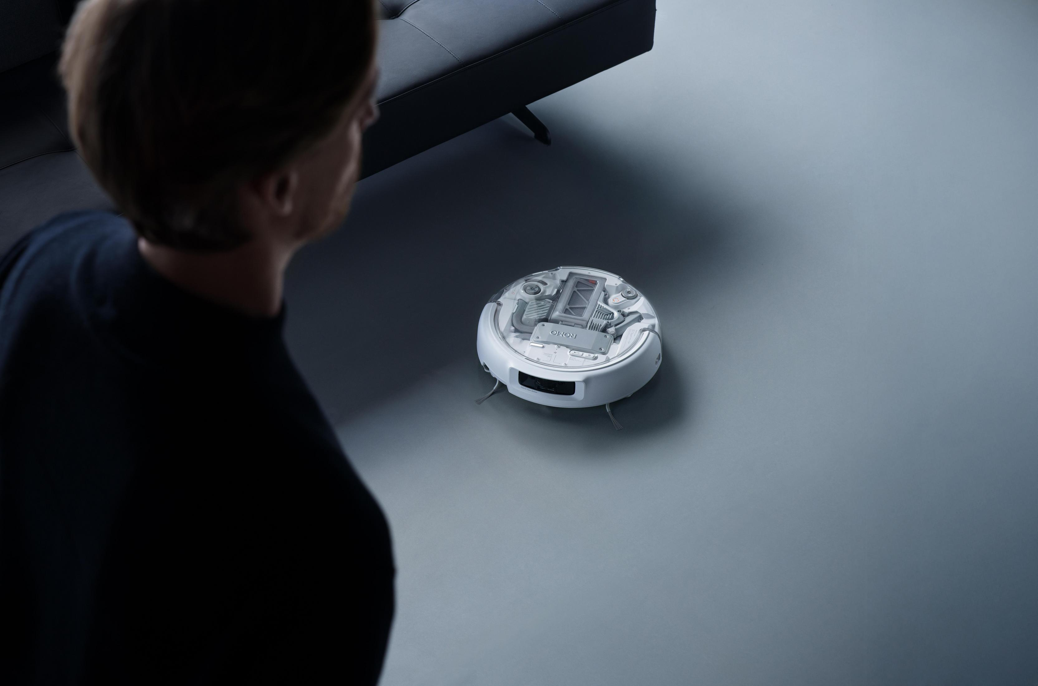 A man looks at a white robot vacuum on a grey floor next to a sofa.