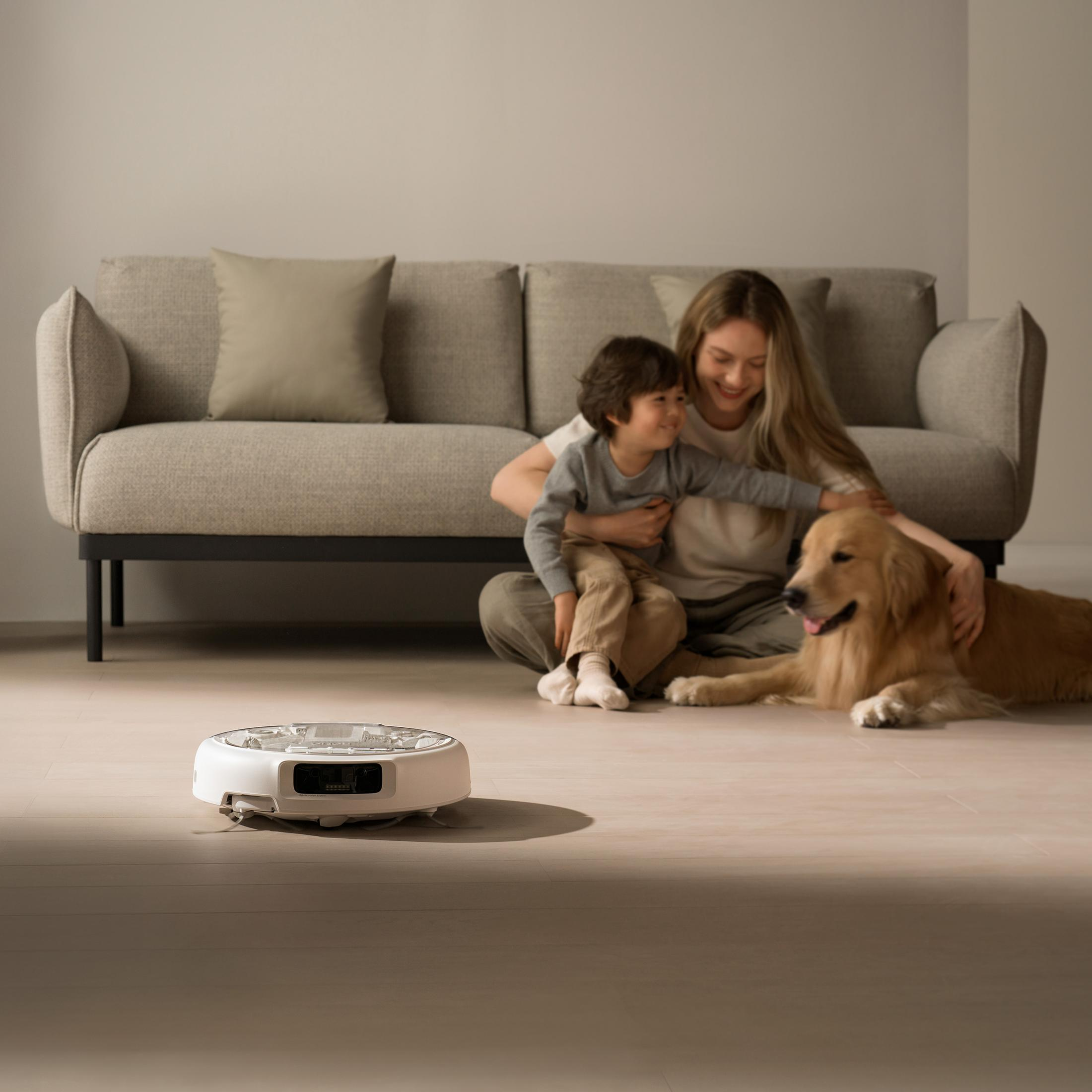 Un robot aspirapolvere bianco su un pavimento in legno chiaro con donna, bambino e cane.