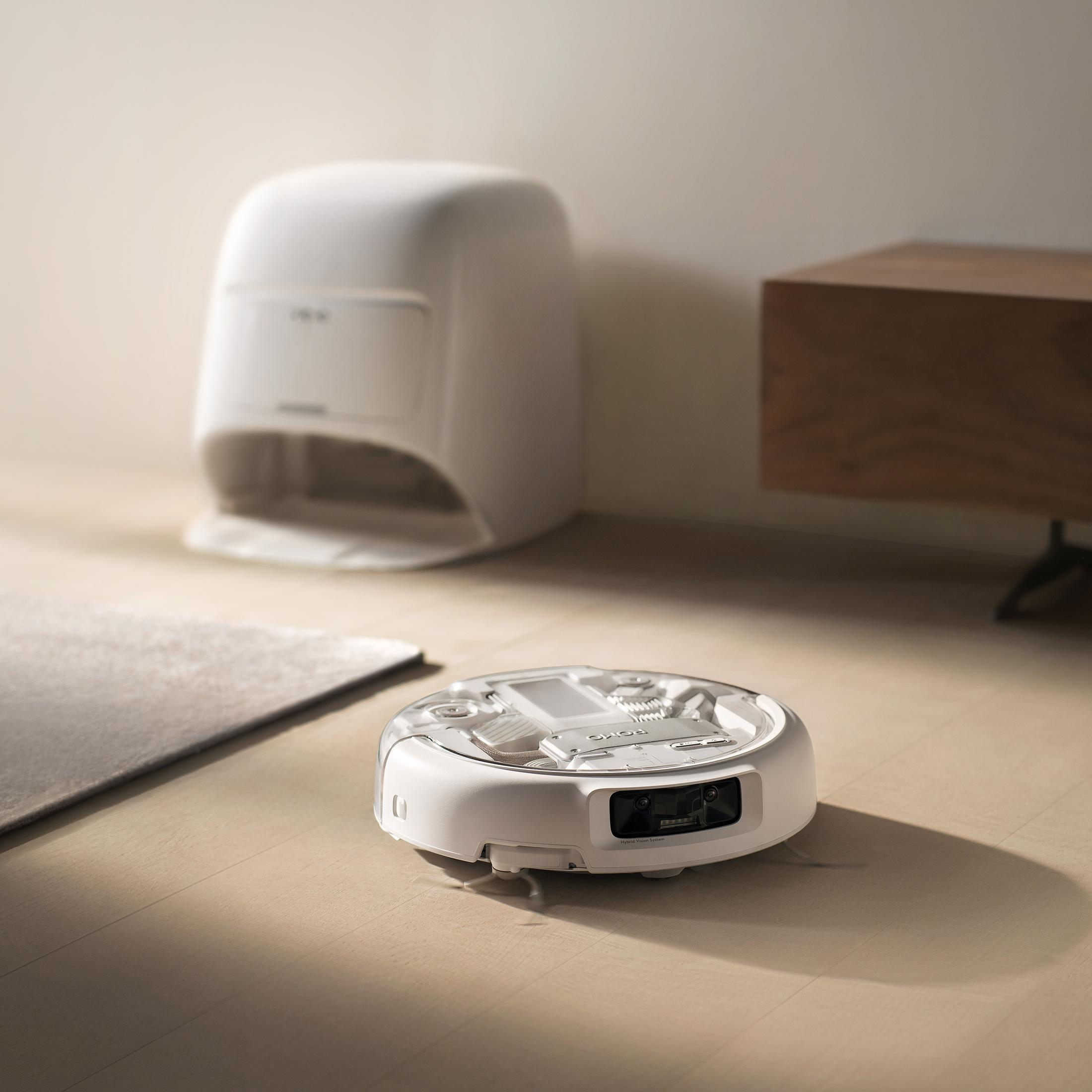 Un robot aspirapolvere bianco con telecamere siede su un pavimento in legno vicino a una base di ricarica e mobili.