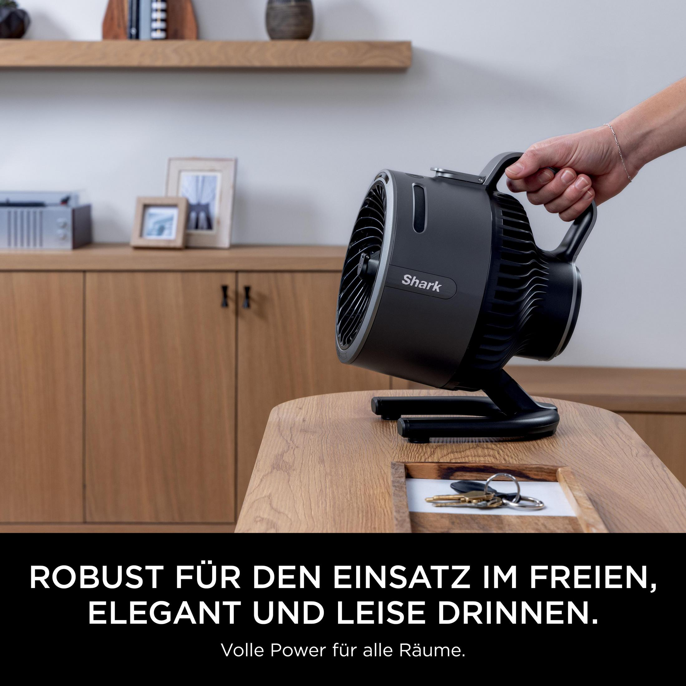 Een hand houdt een zwarte Shark ventilator vast op een houten tafel. Sleutels liggen ernaast.