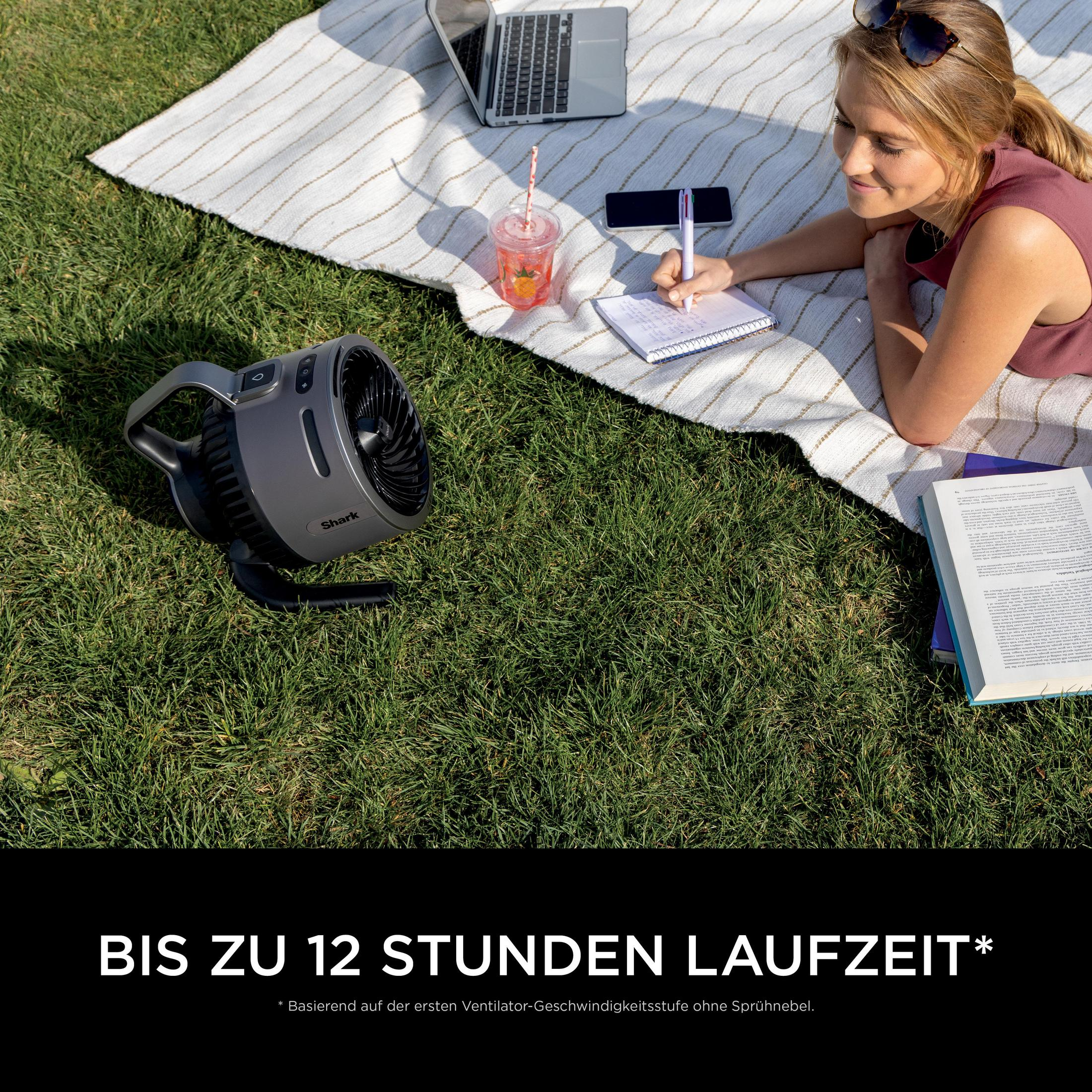 Vrouw ligt op deken in gras te studeren. Ventilator, laptop, drankje, boek.