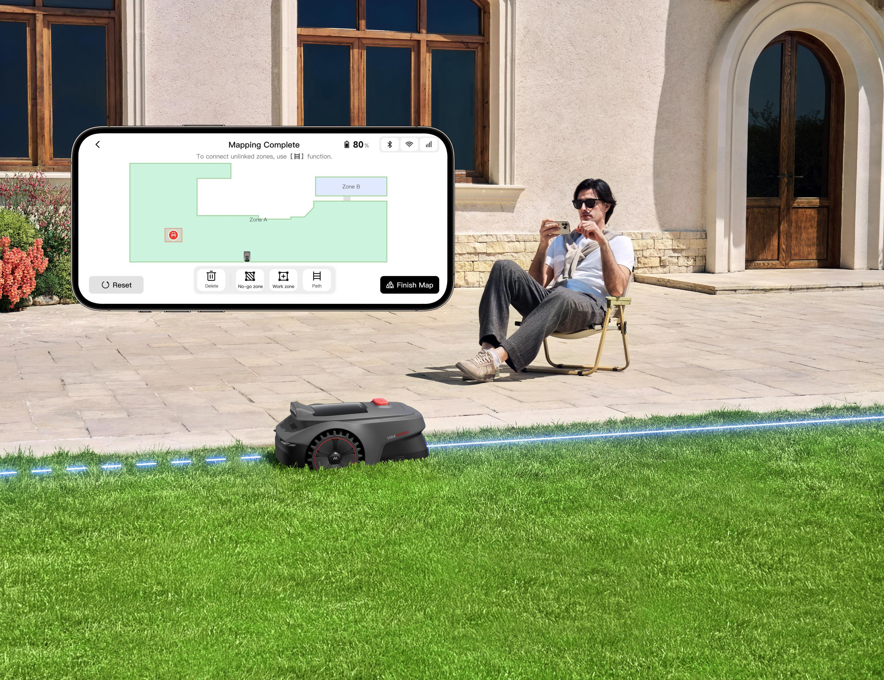 Ein Mann sitzt im Freien und schaut auf sein Handy, während ein Mähroboter auf dem Rasen steht.