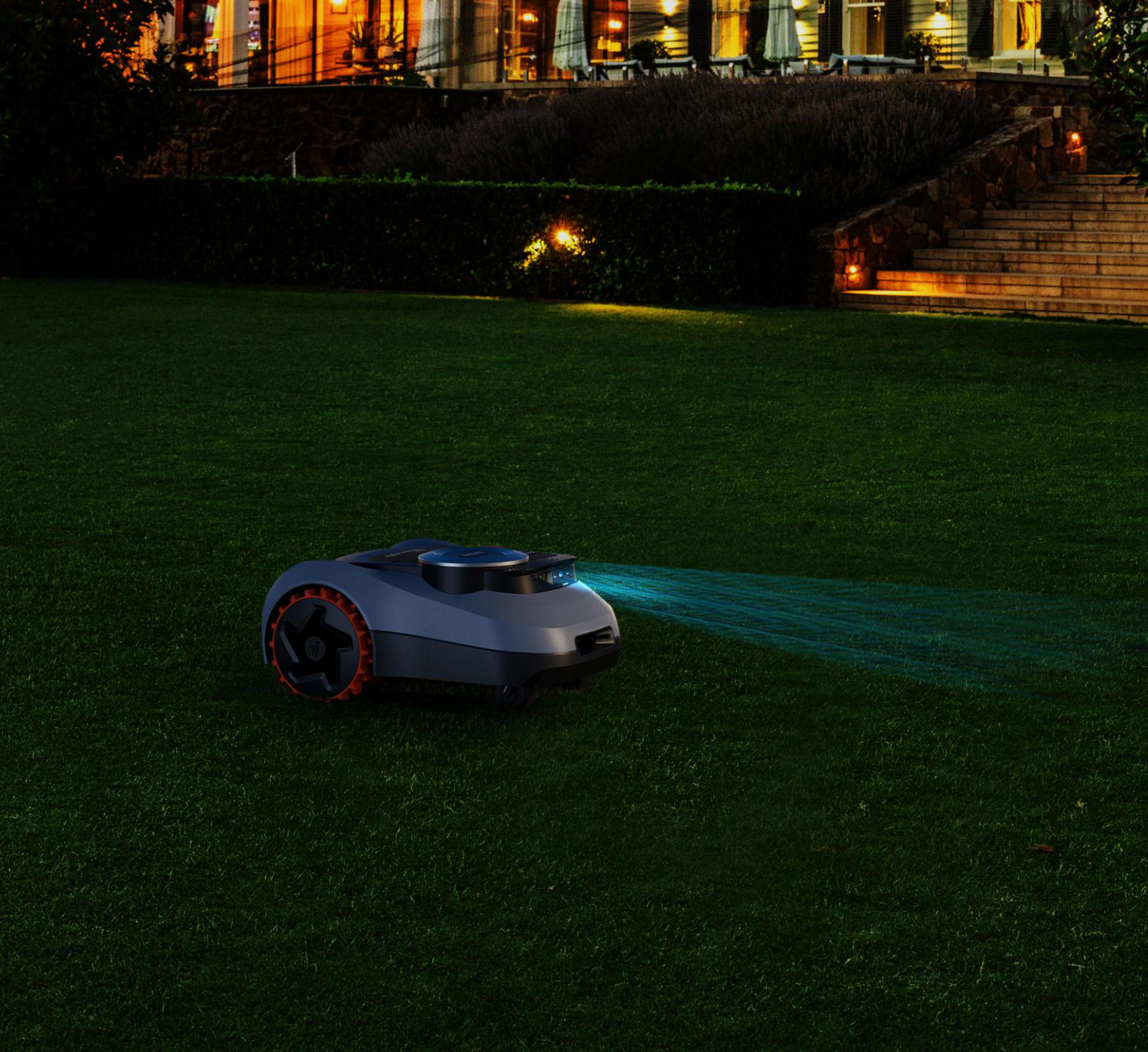 Een robotgrasmaaier maait 's nachts op een donker gazon, met een huis op de achtergrond.