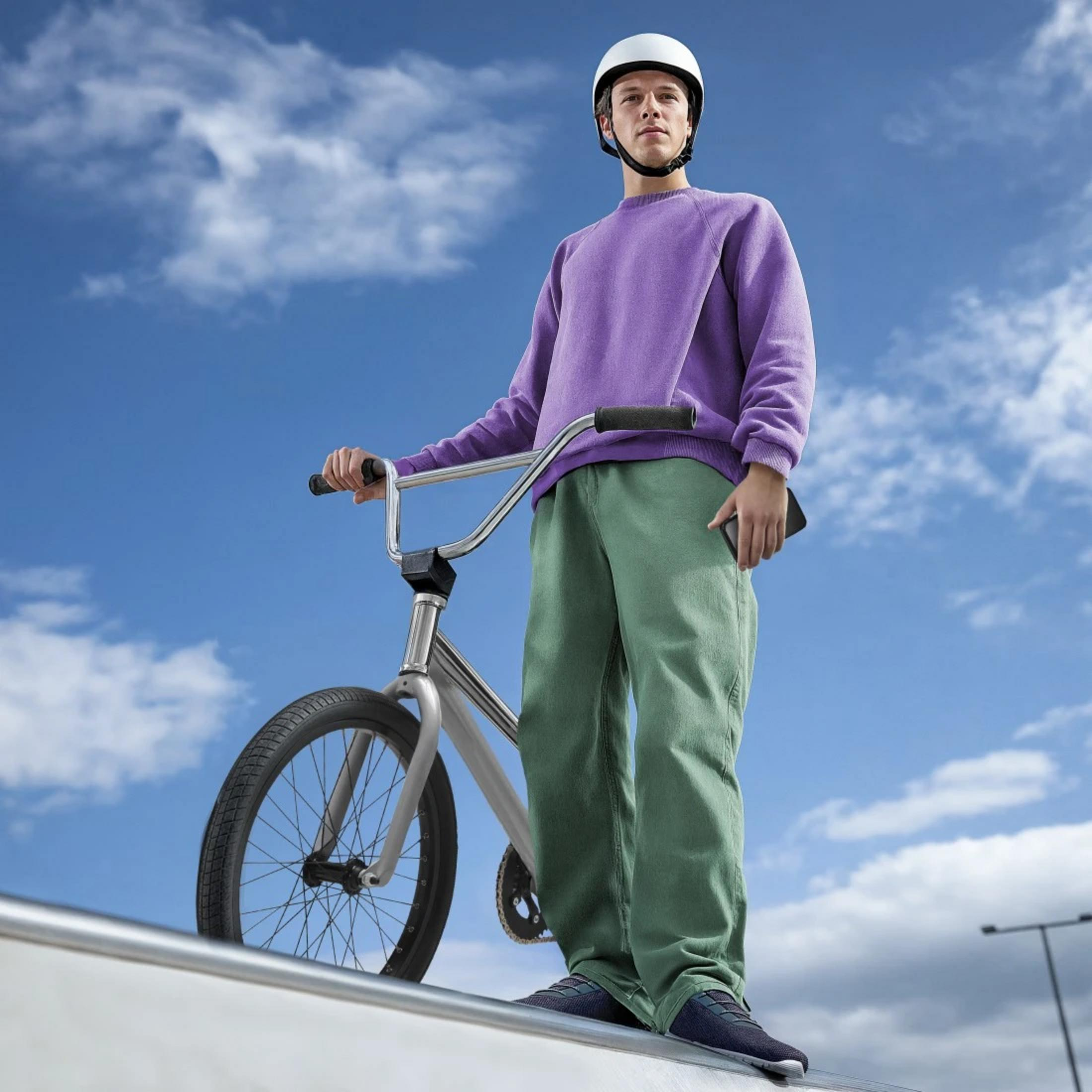 Ein Mann in einem lila Pullover und grüner Hose steht mit einem BMX-Rad auf einer Rampe.
