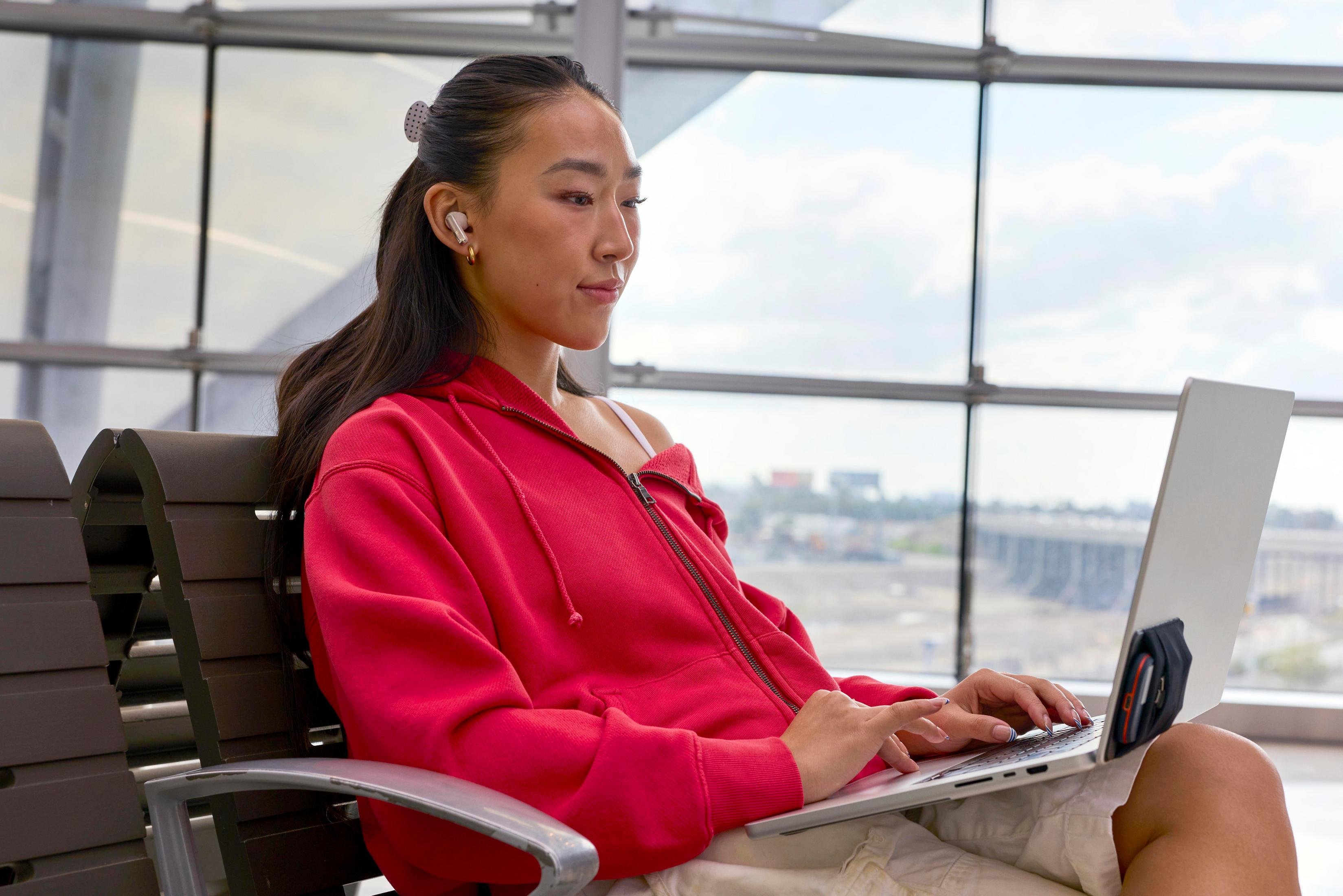 Eine Frau mit Kopfhörern tippt auf einem Laptop, während sie in einem Flughafen wartet.