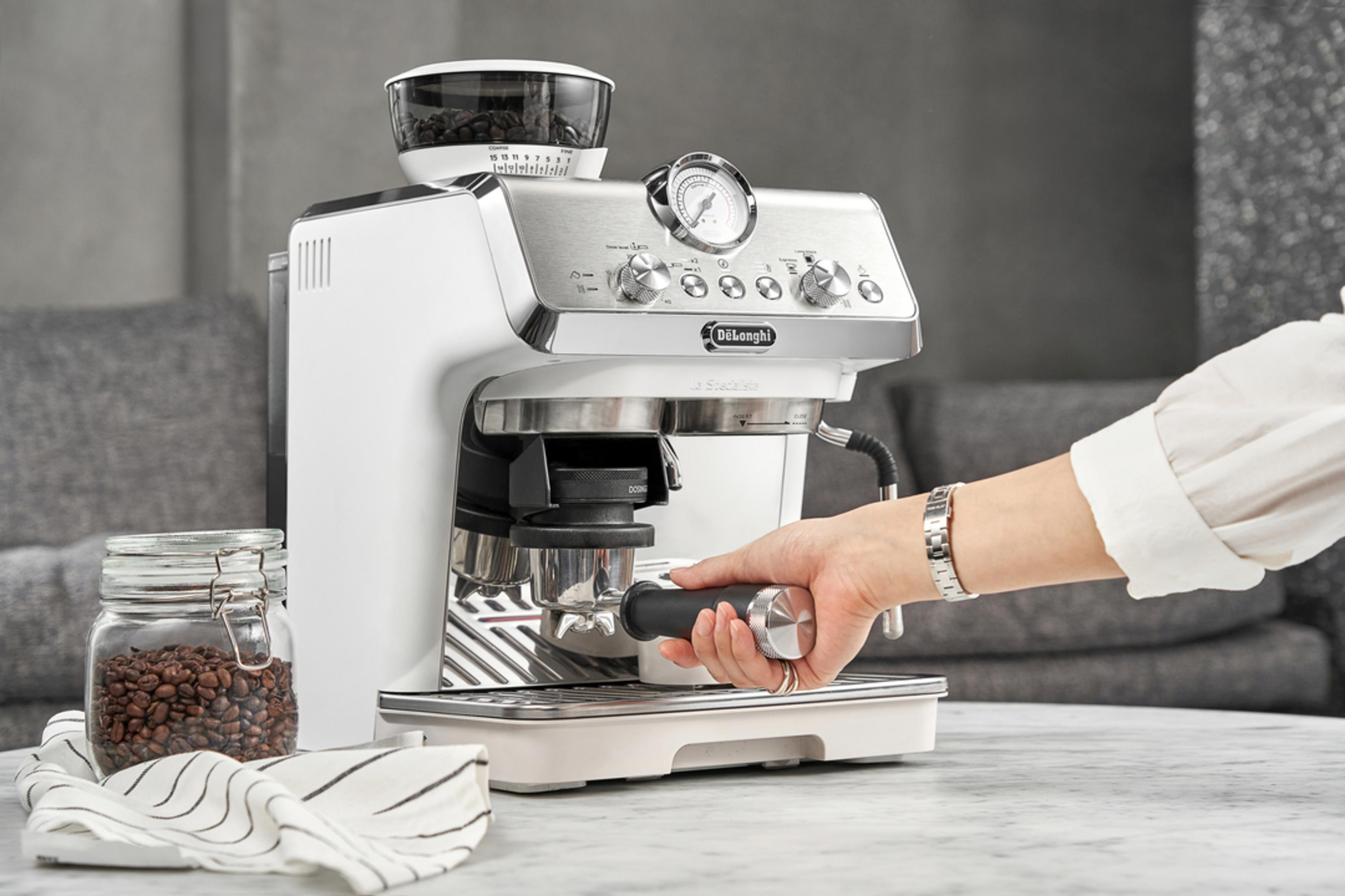 Een hand bedient een witte espressomachine met een pot koffiebonen ernaast.