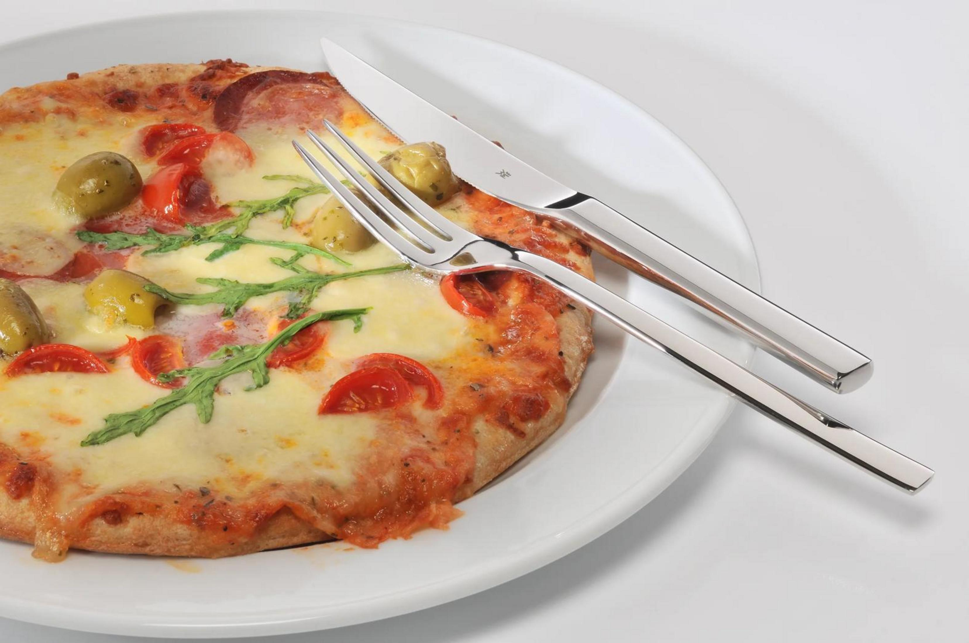 Eine Pizza mit Oliven, Tomaten und Rucola liegt auf einem Teller neben Besteck.