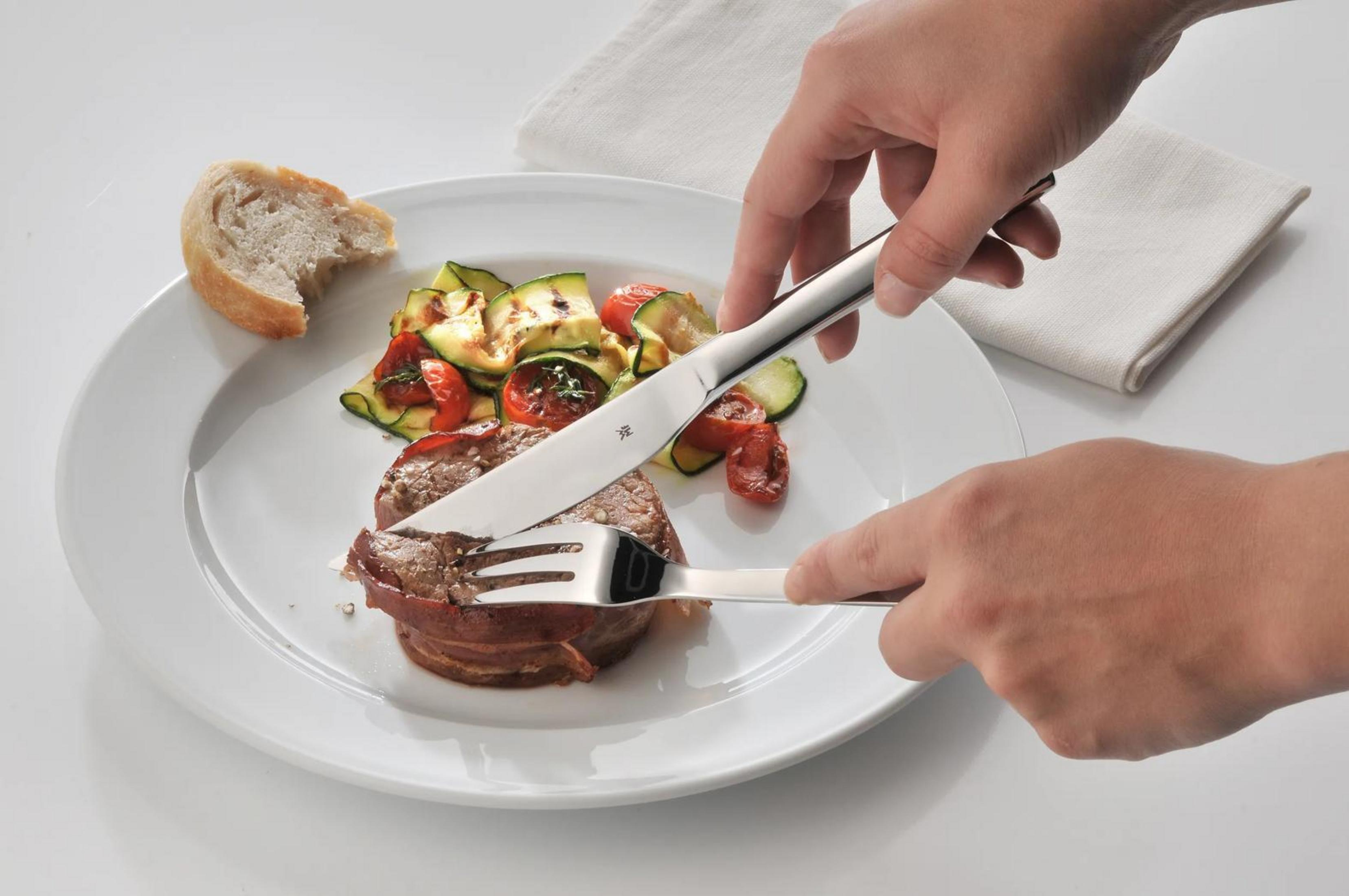 Eine Hand schneidet mit einem Messer in ein Rinderfilet, daneben gegrilltes Gemüse und Brot.