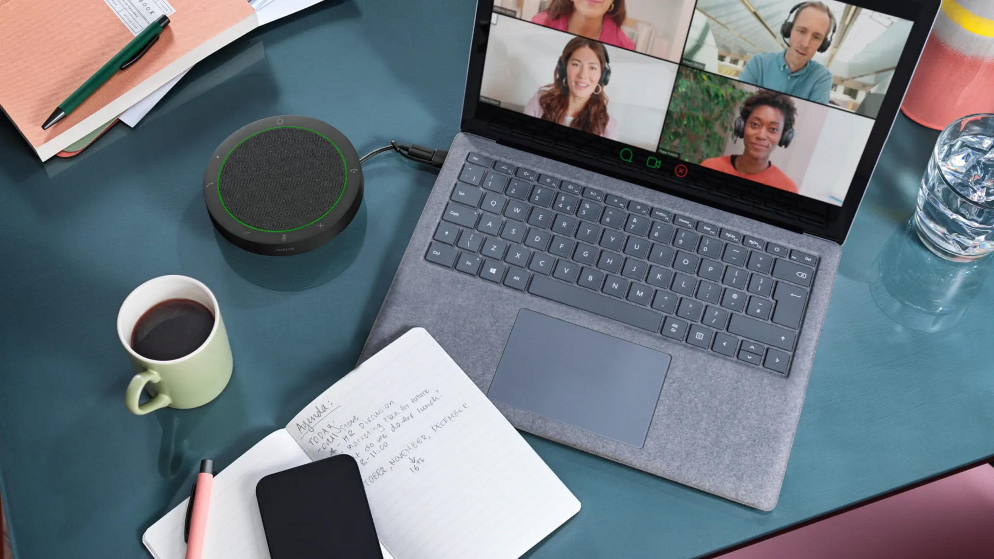 Laptop mit Videokonferenz, Lautsprecher, Kaffee, Notizbuch und Stift auf Schreibtisch.