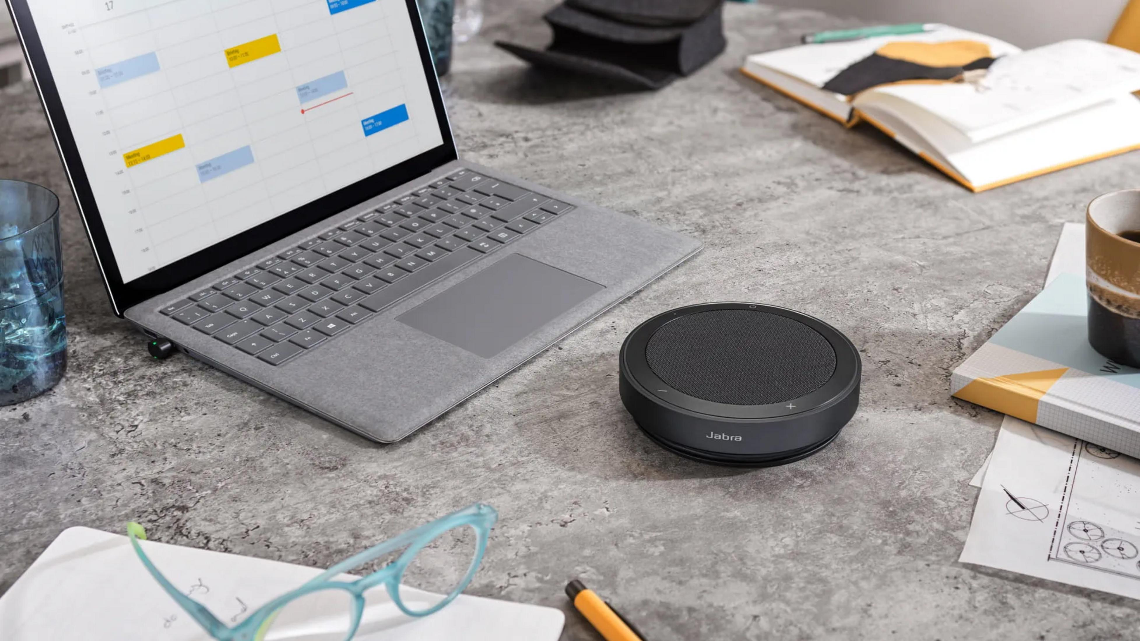 Un laptop, altoparlante Jabra, occhiali e caffè su una scrivania.