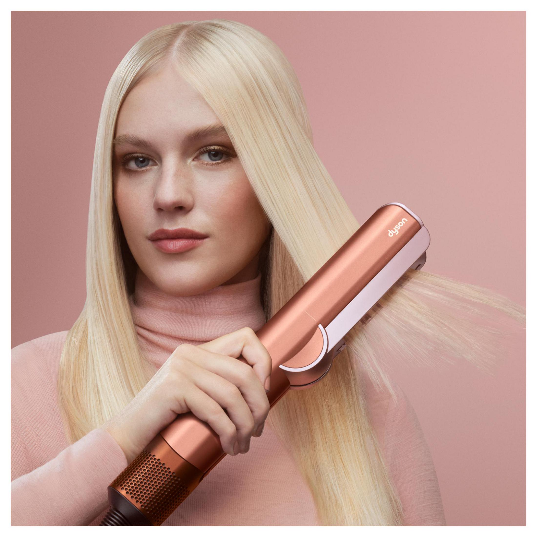 Frau mit blonden Haaren stylt ihr Haar mit einem roségoldenen Dyson-Haarglätter vor einem rosafarbenen Hintergrund.