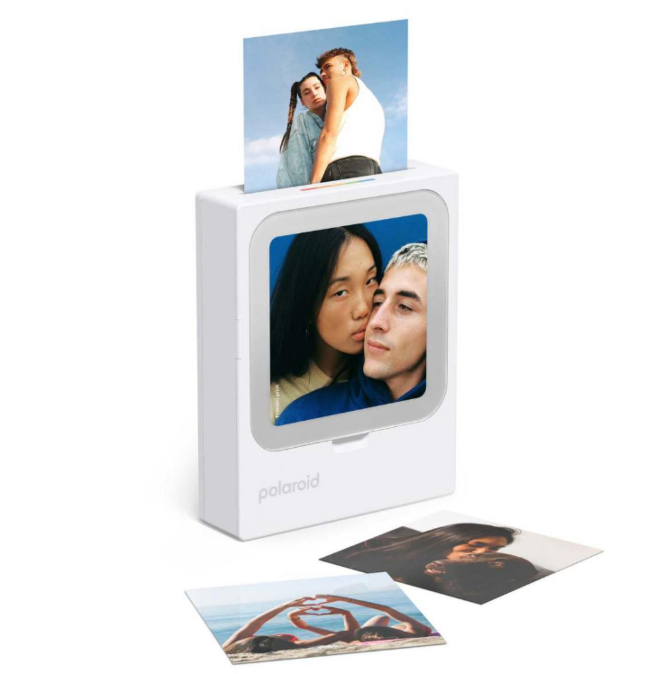 Ein weißer Polaroid-Drucker gibt zwei Fotos aus. Unten liegen drei weitere Fotos.