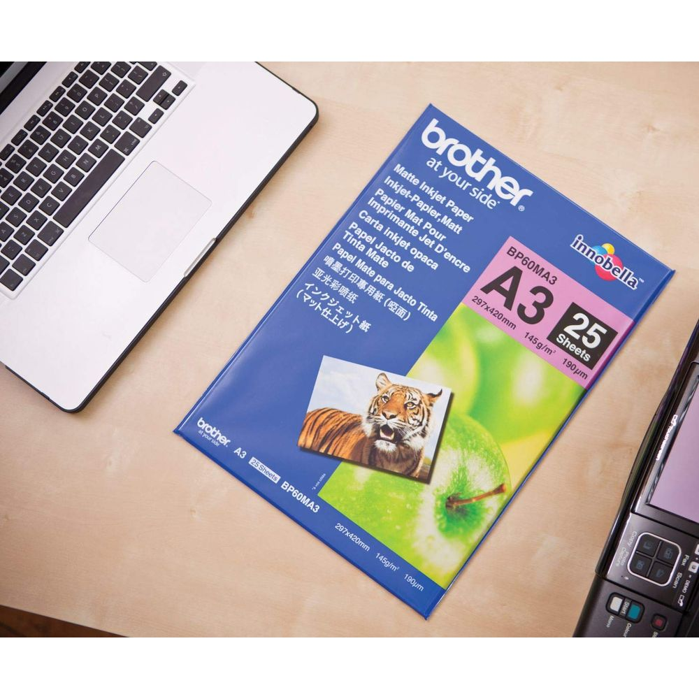 Ein Laptop, Brother A3 Matte Inkjet Paper und ein Handy liegen auf einem Holztisch.
