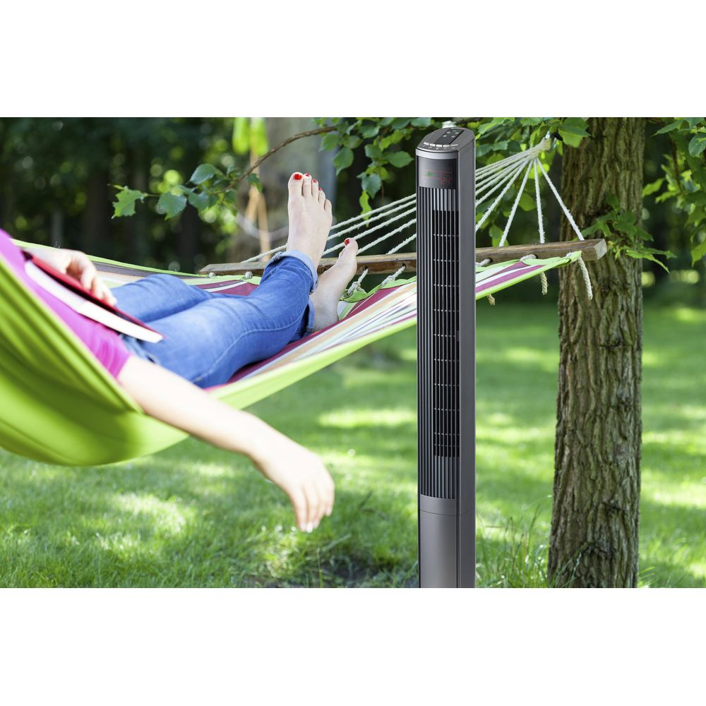 Eine Person liegt in einer Hängematte im Garten und liest ein Buch. Ein Turmventilator steht daneben.