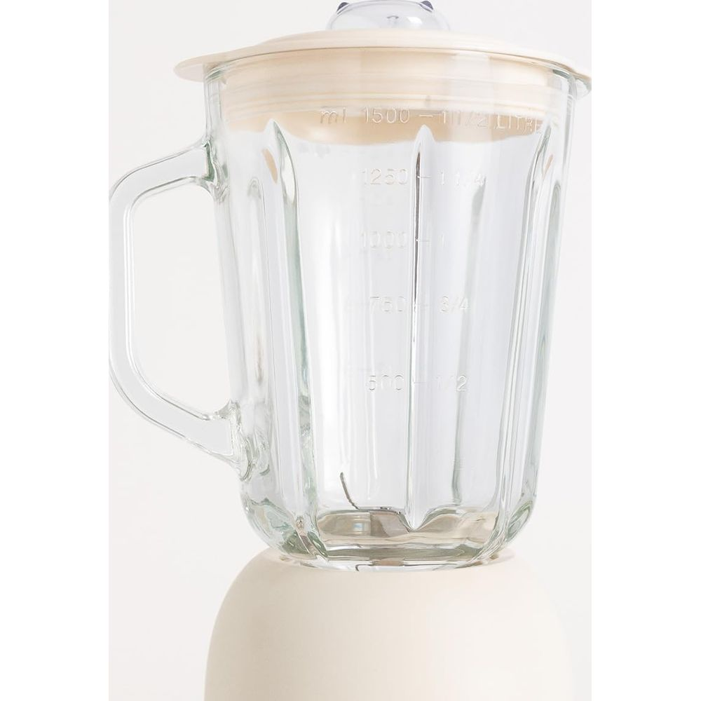 Un vaso de licuadora de vidrio con asa y marcas de medida, sobre una base crema.
