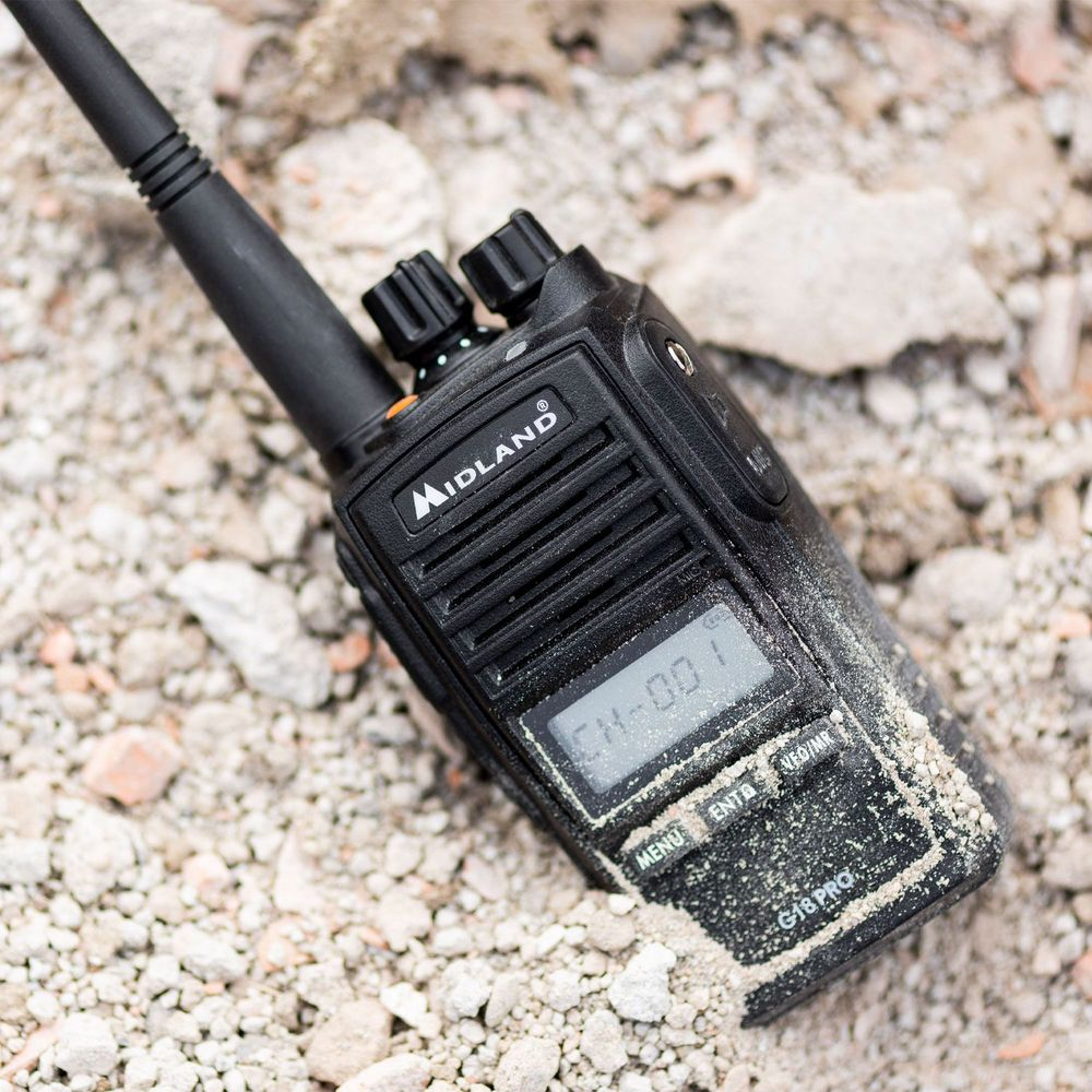 Un walkie-talkie Midland negro yace sobre escombros grises, cubierto de arena.