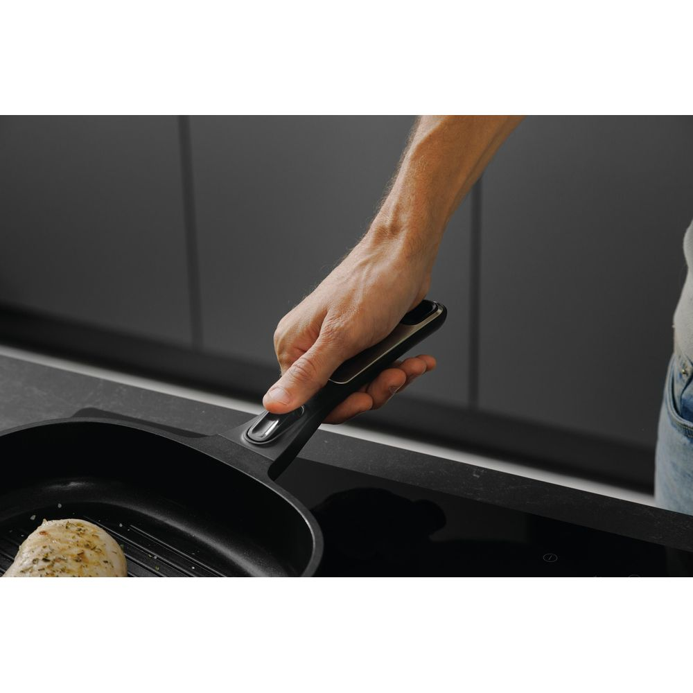 A hand holds a grill pan handle over chicken in a pan on an induction hob.